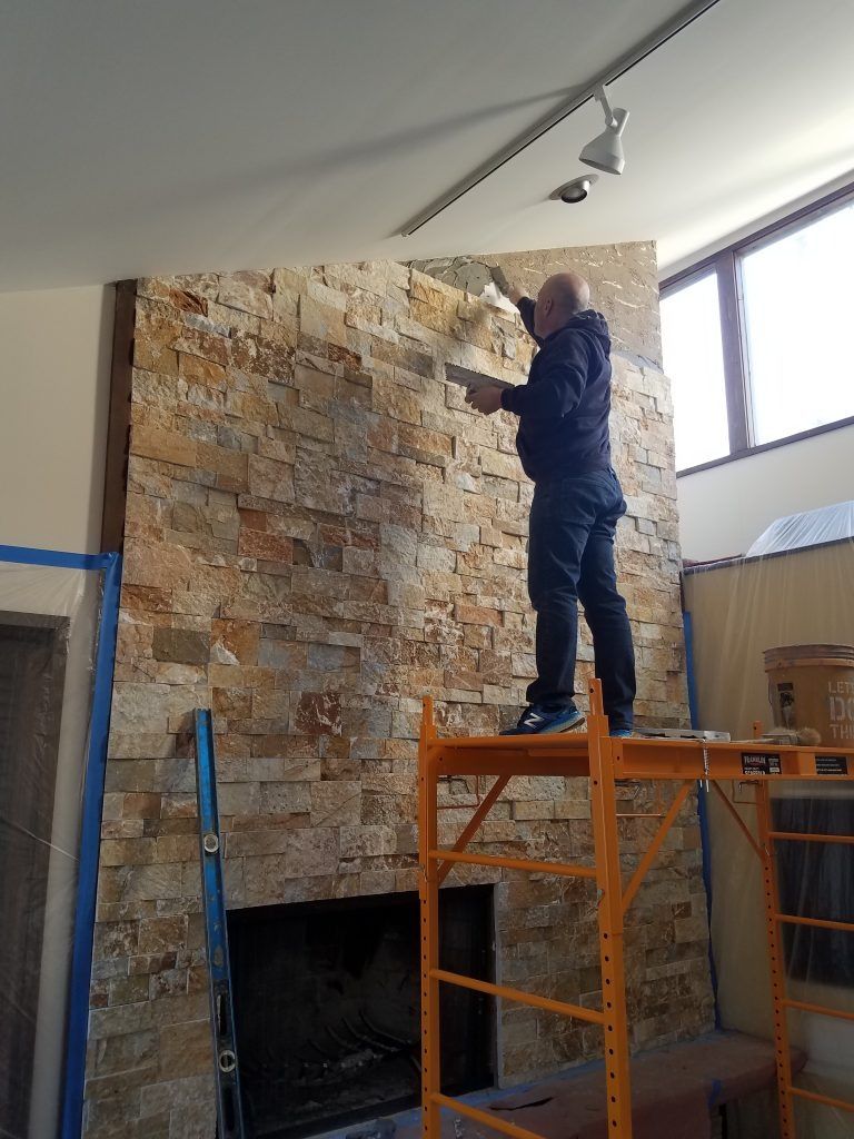 Man on a scaffold applying grout to a stone fireplace. Interior shot with windows, tan and brown stone.
