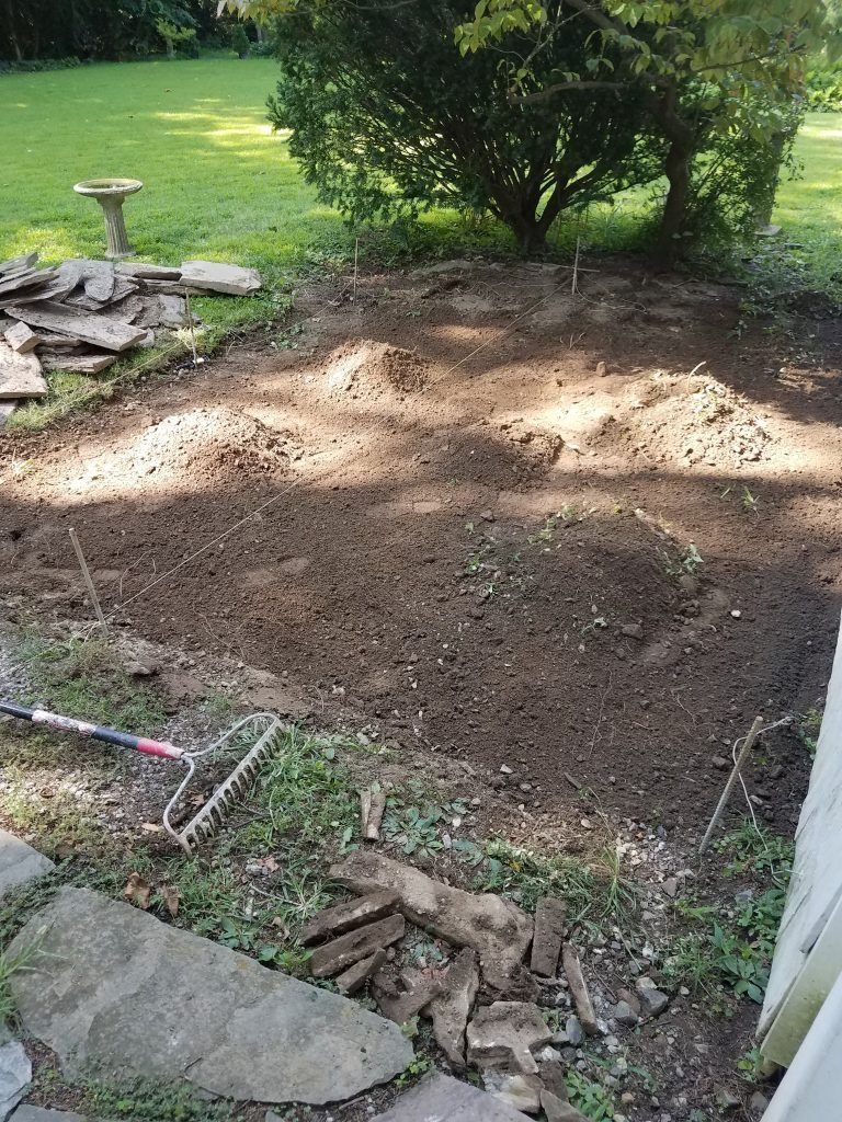 A dirt patch in a yard, prepared for planting; surrounded by grass, stones, and a tree in the background.