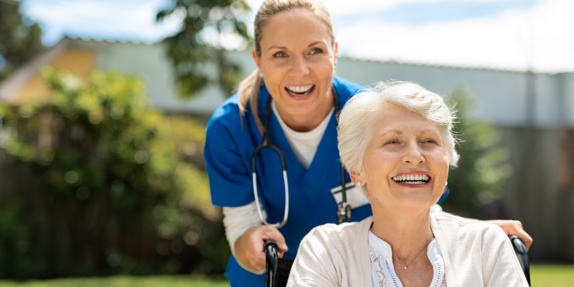 A healthcare provider and an older adult laugh while looking at a tablet together on a couch.