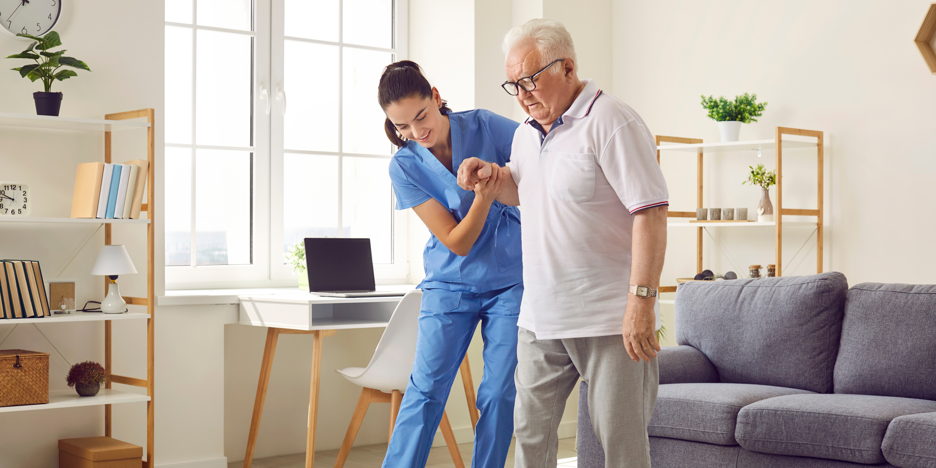 A caregiver in blue scrubs assists an older person in a white polo shirt as they stand up in a bright, modern living room.