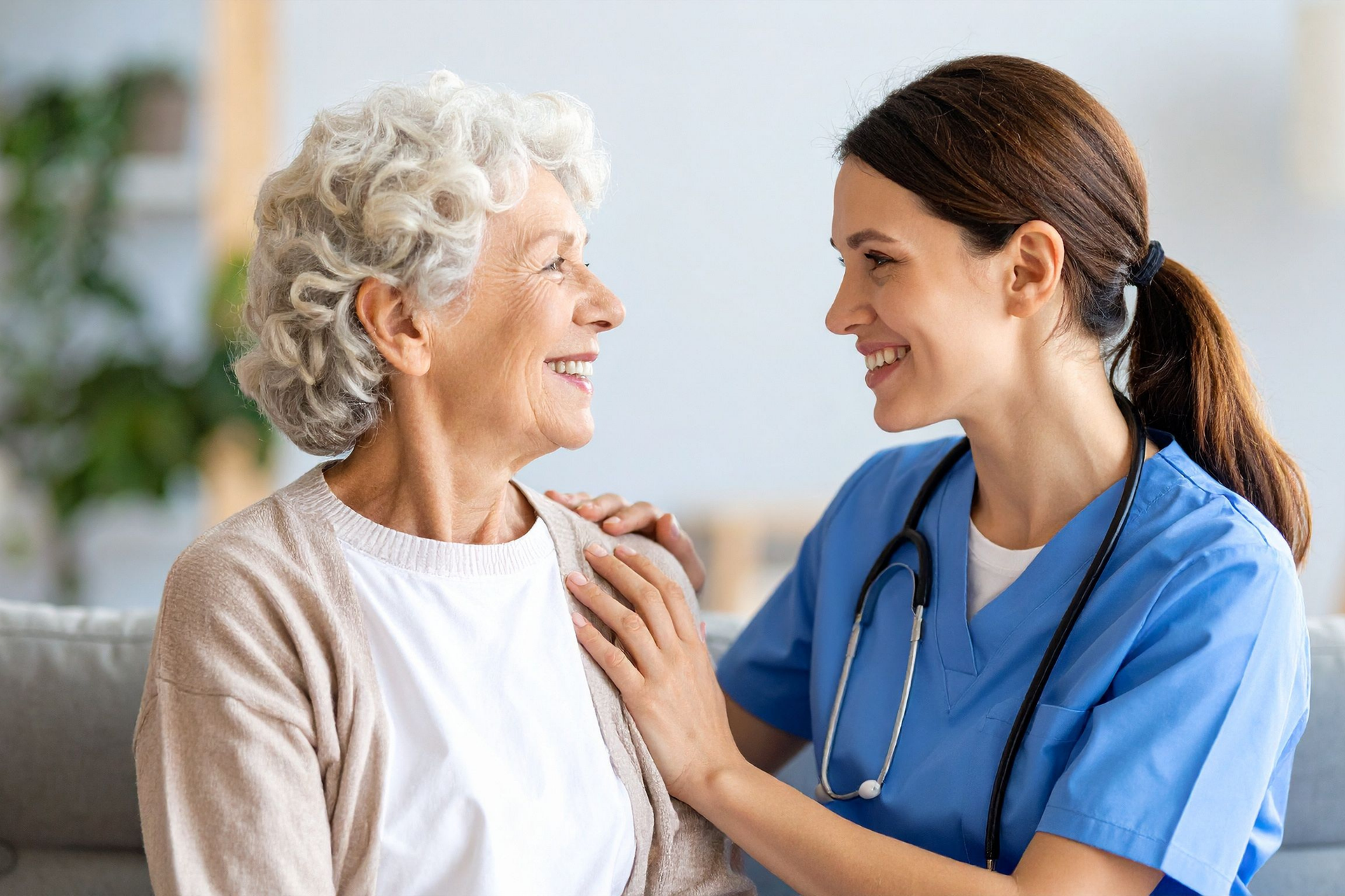Nurse assists patient using a walker in a home. The patient wears glasses and pink shirt. The nurse smiles.