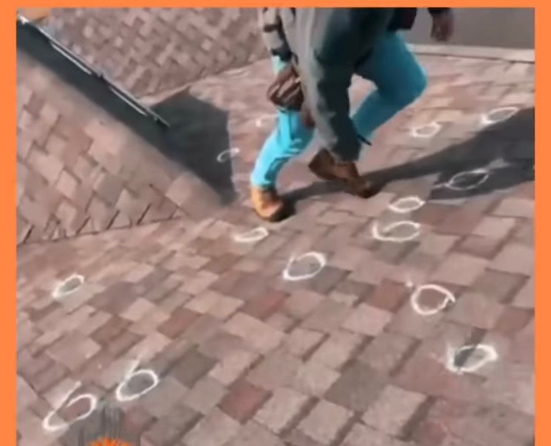 Person walking on a roof, marking hail damage with white circles. Brown shingles, blue pants, and work boots are visible.