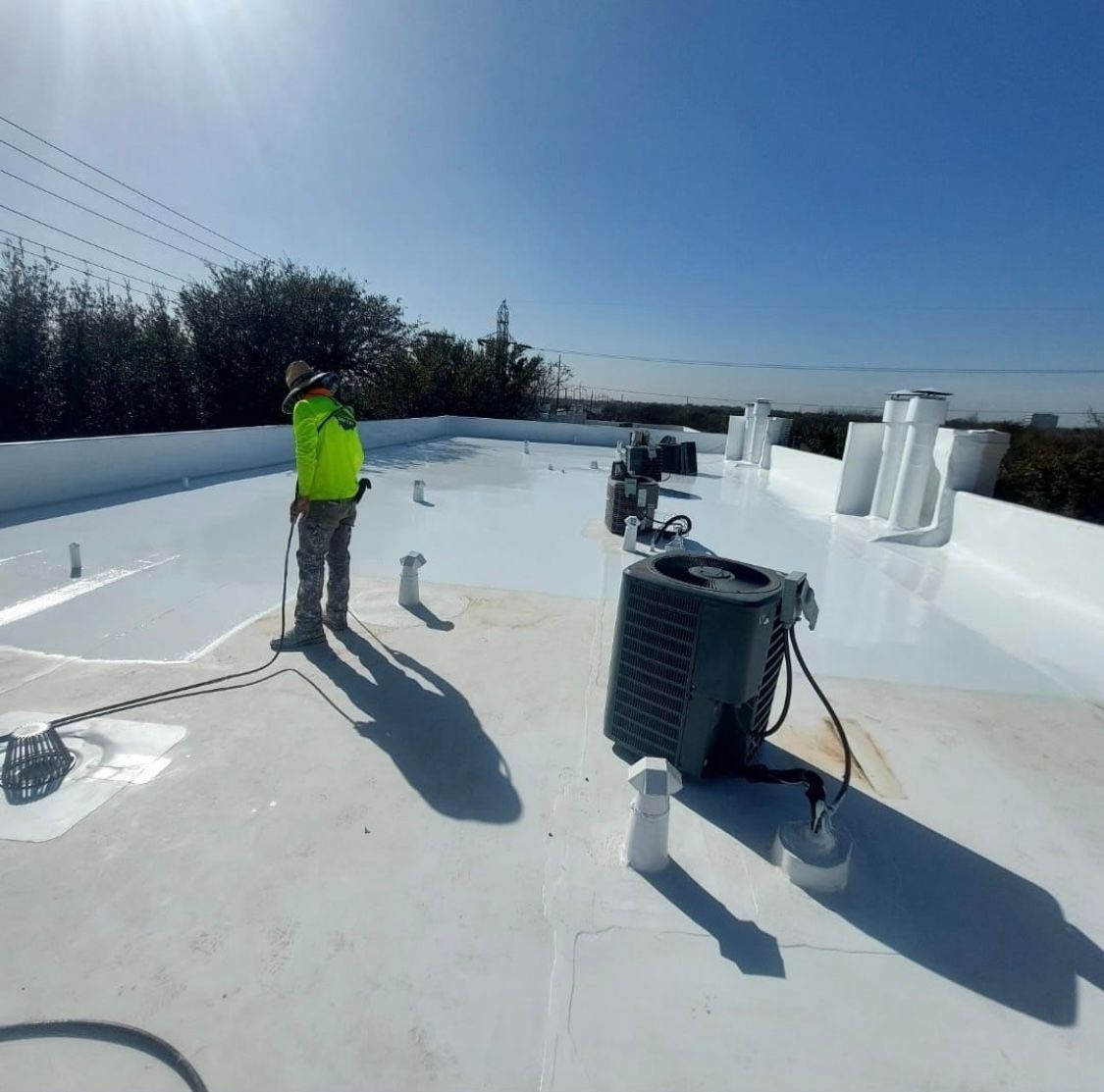 A worker in a neon green vest applies white coating to a flat roof under a blue sky.