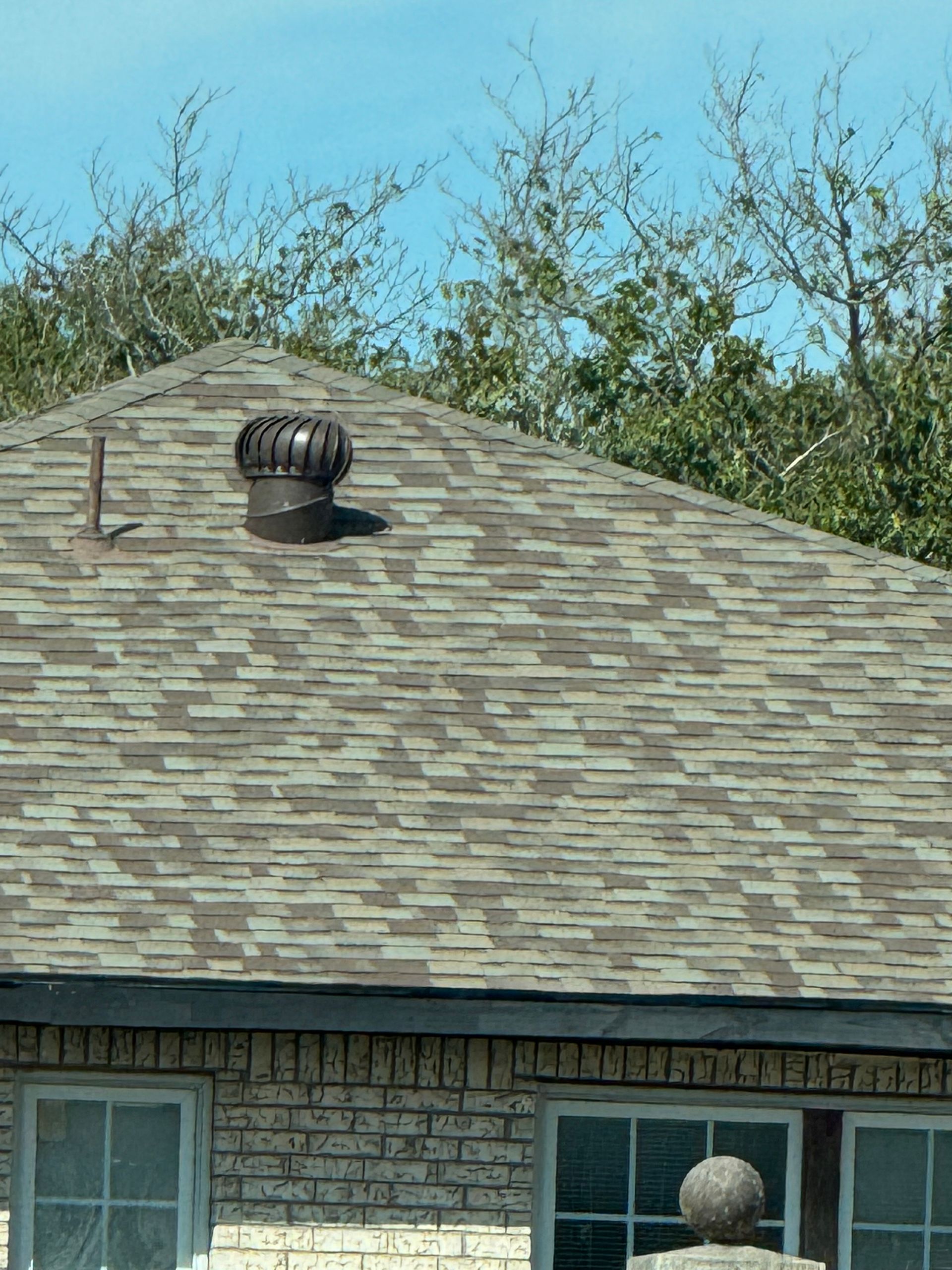 Brown and grey shingle roof with a rotating vent and chimney, set against a blue sky with green trees.