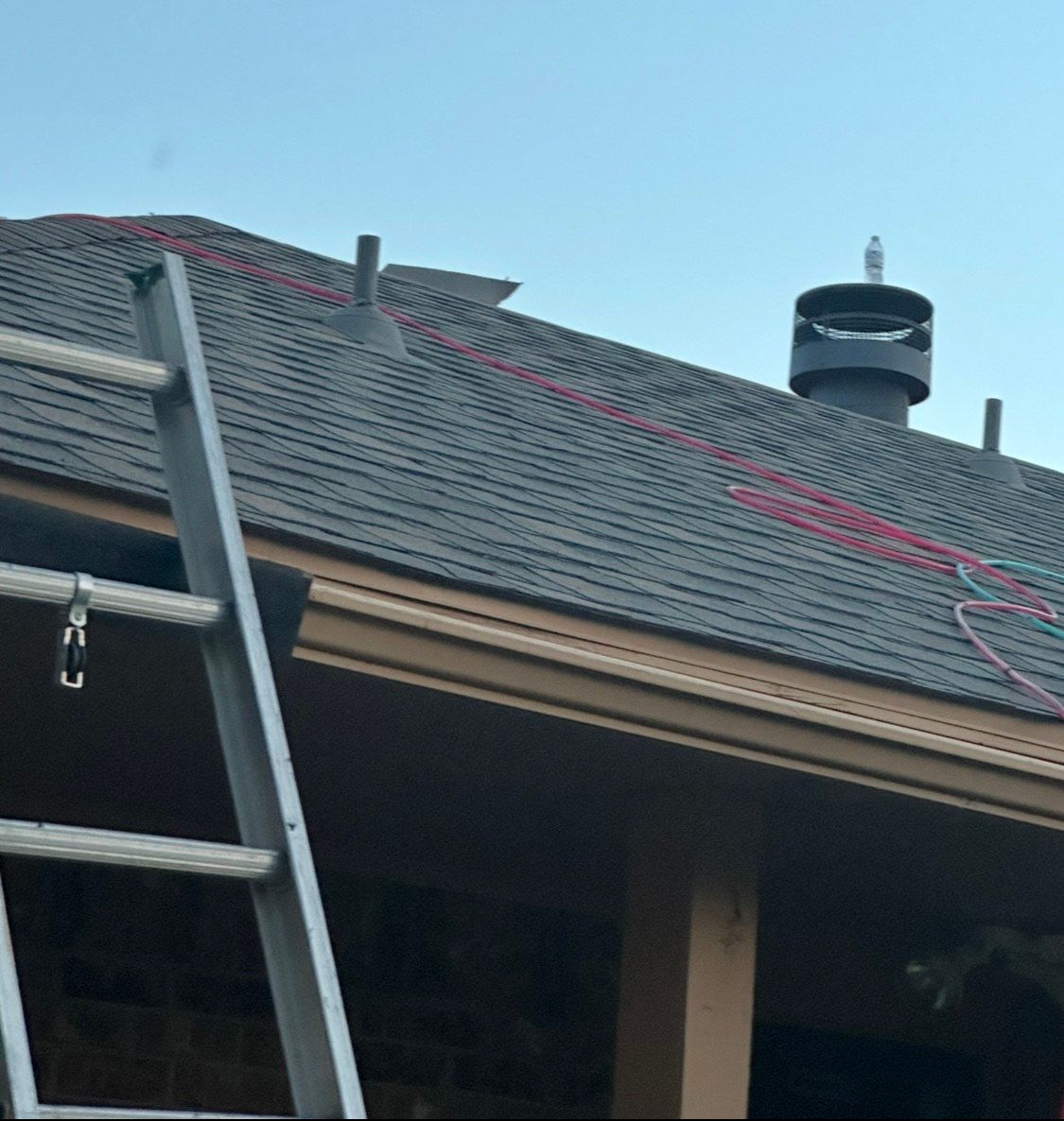 Ladder leaning against a dark shingle roof with a chimney and red cable; clear sky.