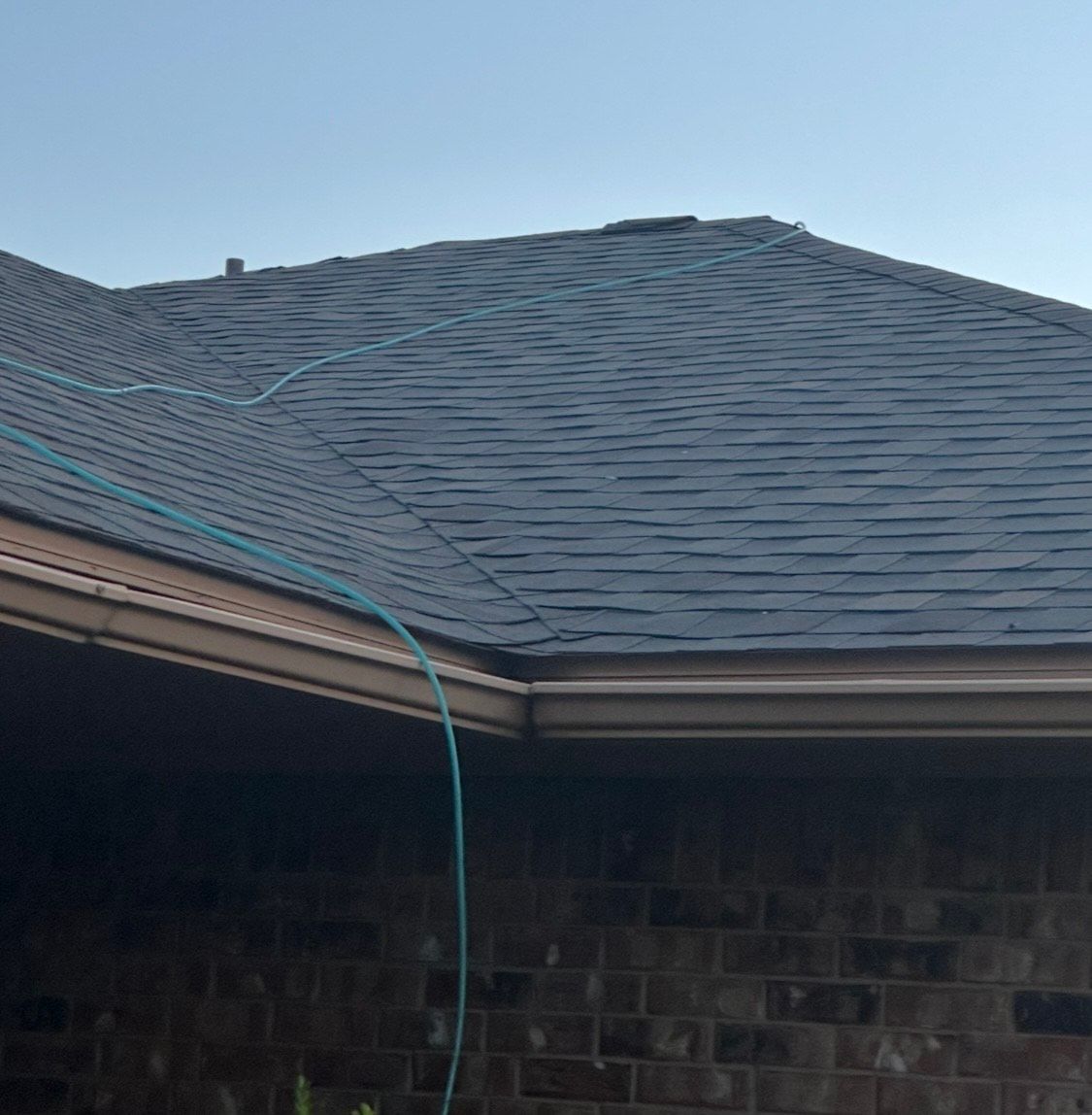 A dark shingle roof with a blue hose over a brick wall and a brown gutter against a blue sky.