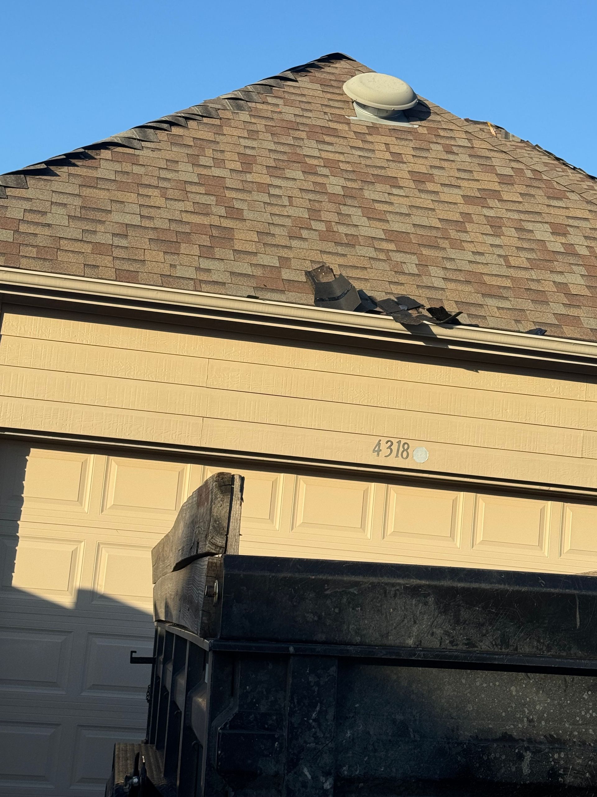 A black dump truck in front of a tan building with a brown shingled roof under a clear, blue sky.
