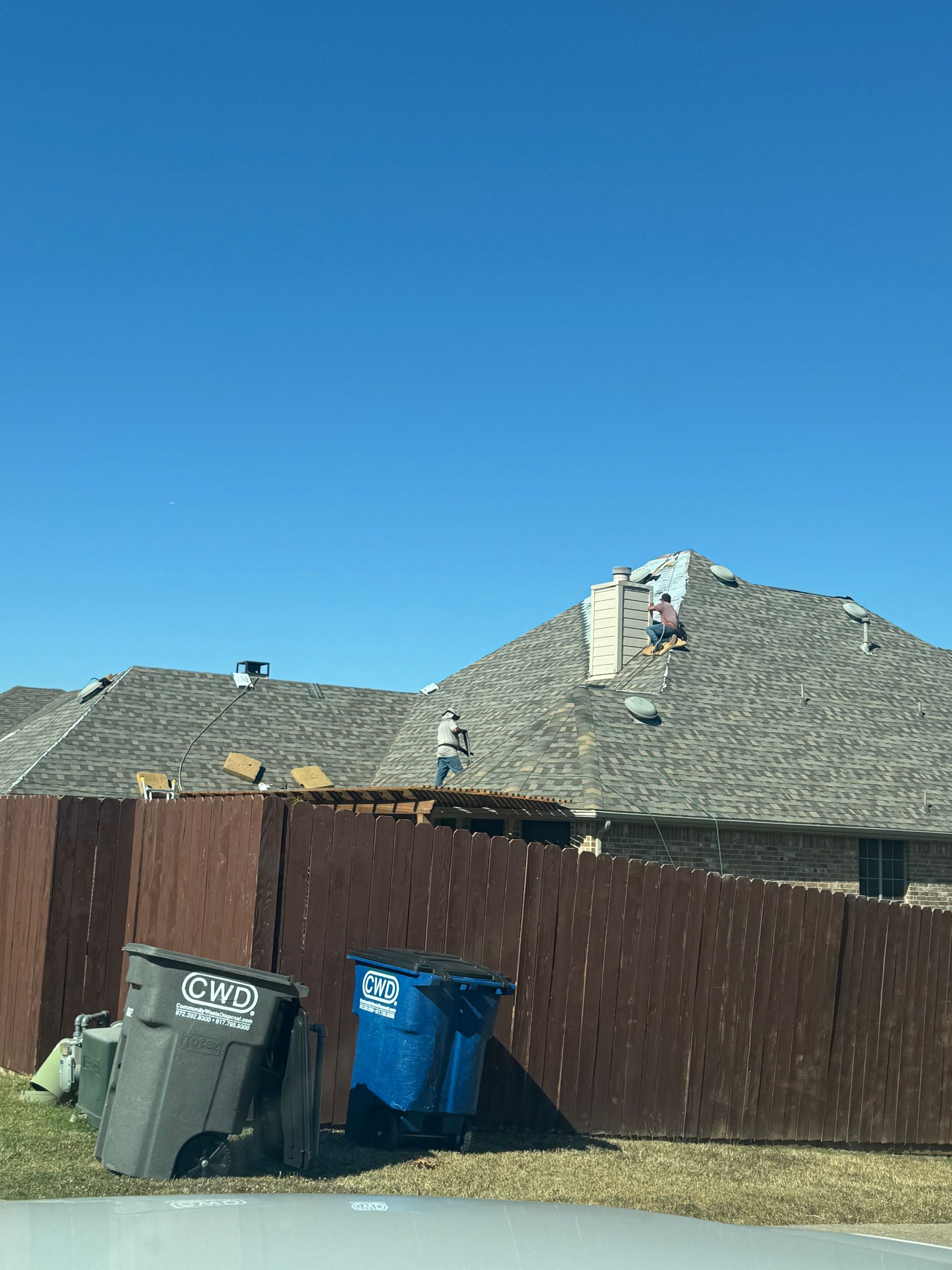 Damaged house roof behind a brown fence, trash cans in the foreground under a clear blue sky.