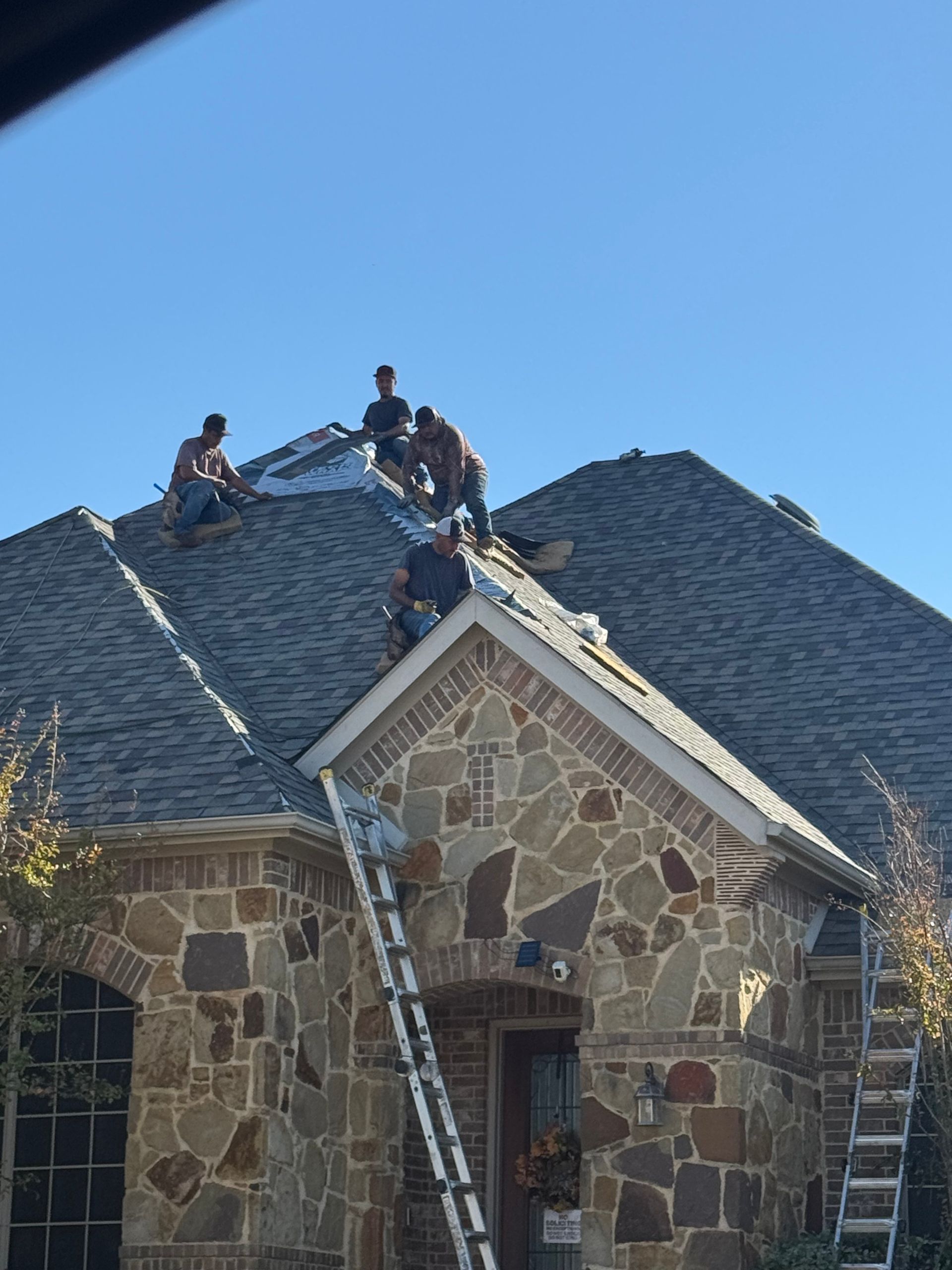 Roofers on a house with stone facade, replacing shingles on a sunny day. Ladders propped up.