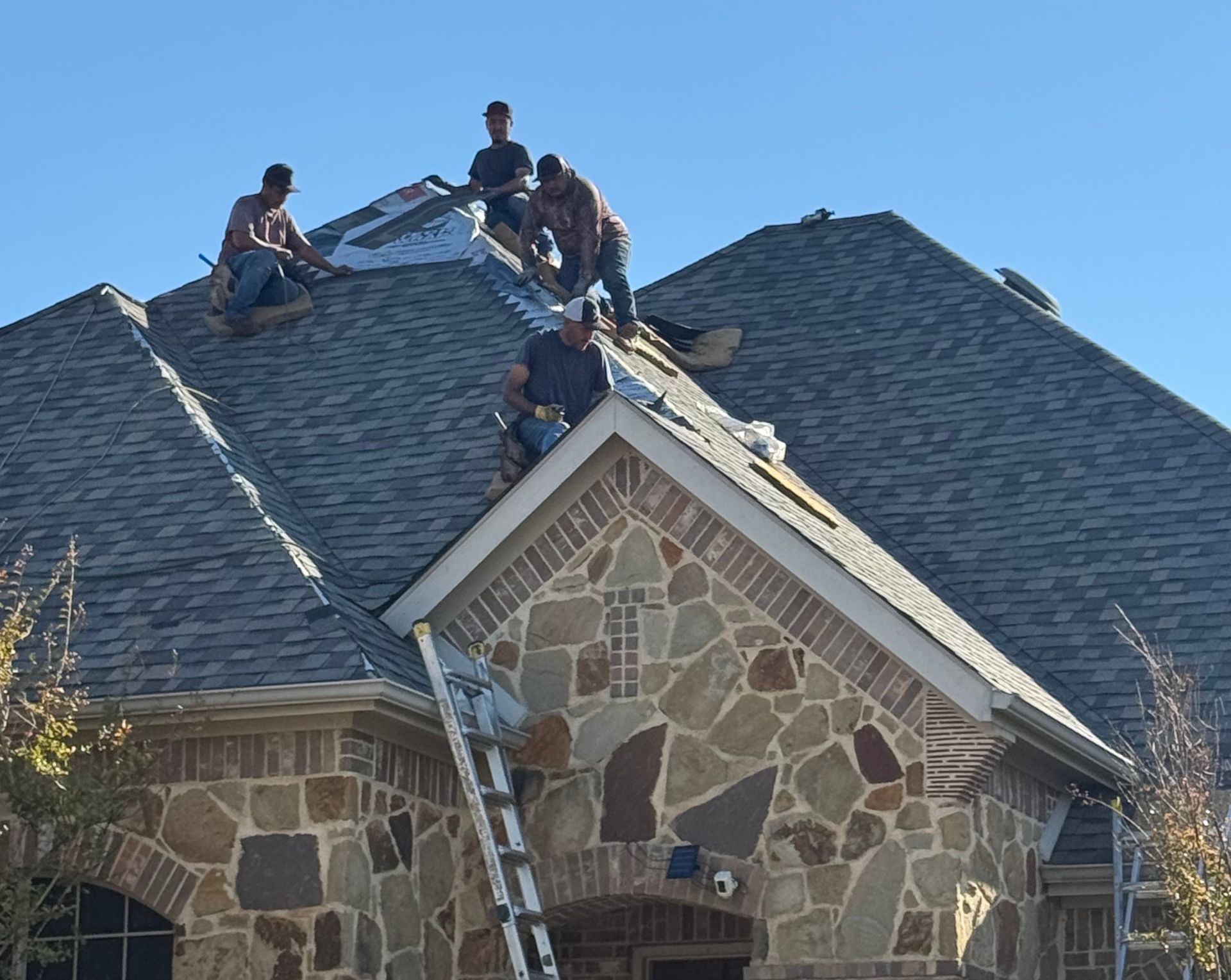 Roofers on a house with stone and brick exterior, replacing roof shingles. Ladder present.
