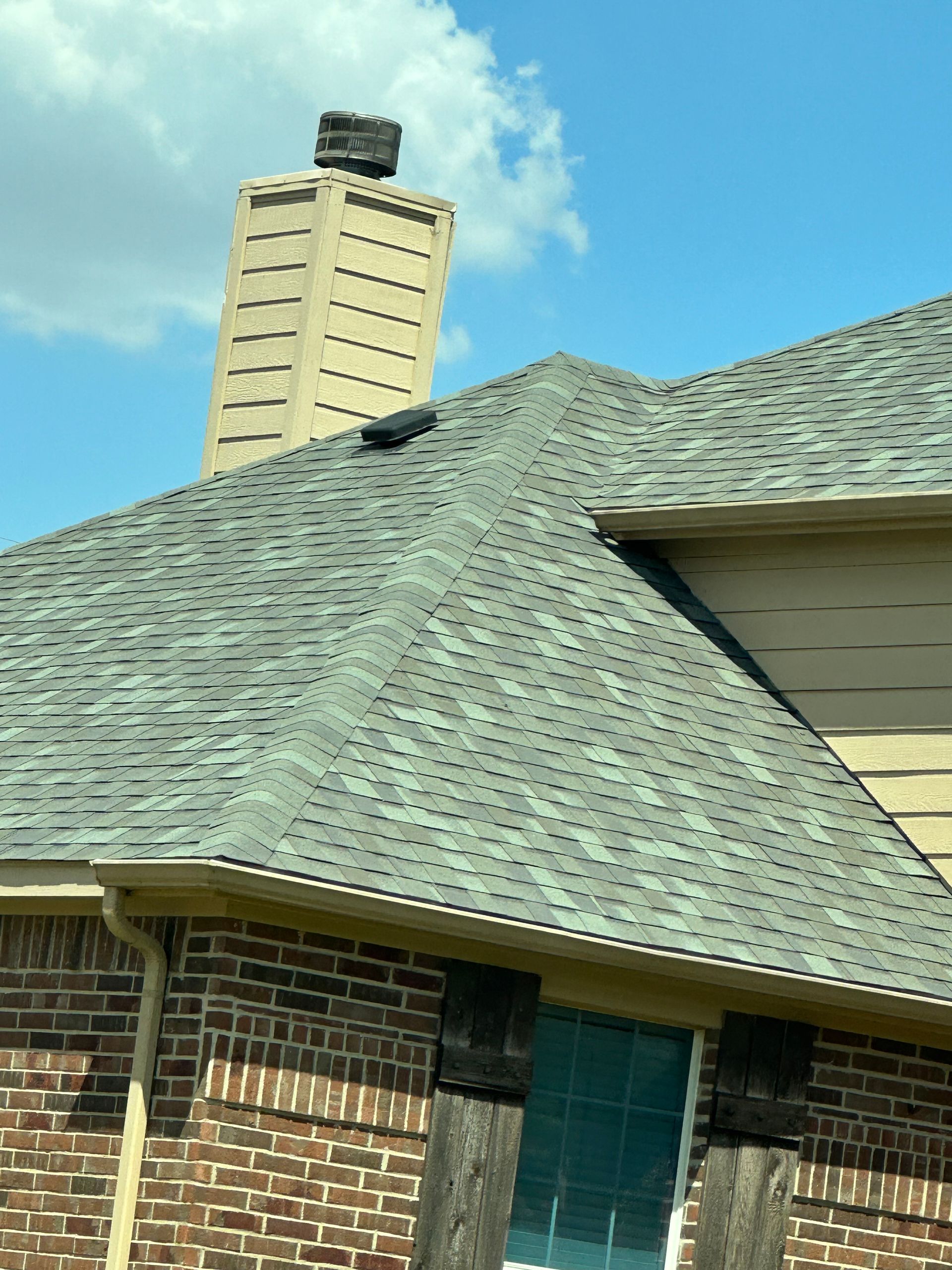 Gray shingled roof with a chimney and part of a brick house against a blue sky.