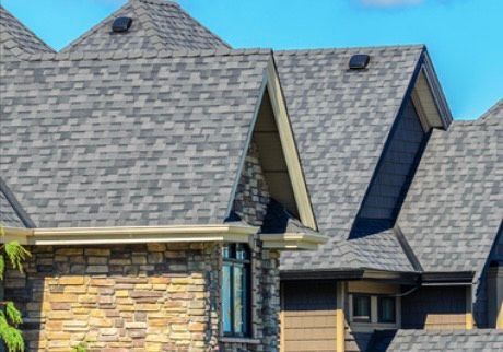 Gray shingle roof on a house with stone and brown siding, against a blue sky.