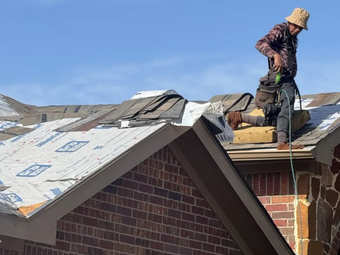 Roofer on a partially torn-off roof, working under a clear blue sky. Brick home in foreground.