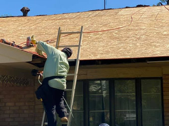 Person on a ladder working on a roof, securing something. House visible with visible plywood and orange wire.