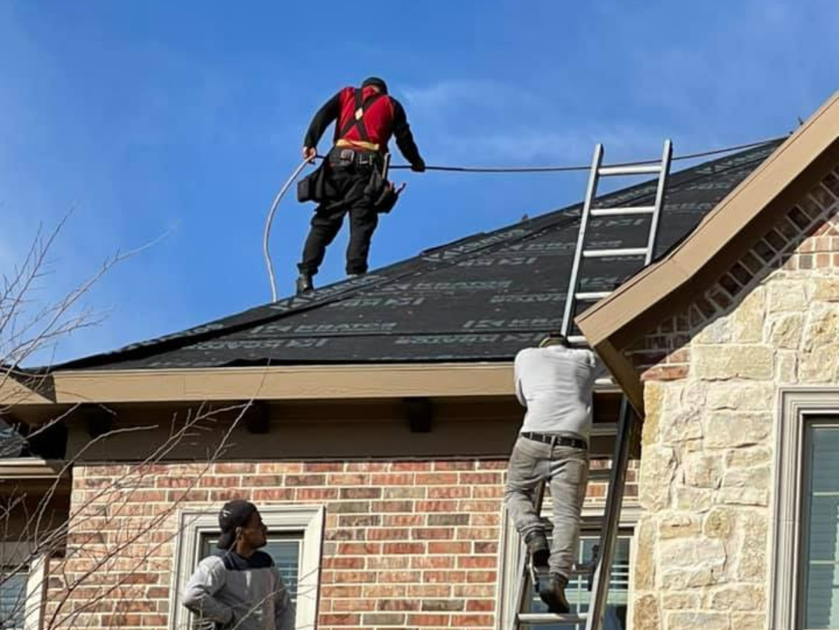 Roofers working on a residential roof, one on the roof, two on a ladder and ground, brick and stone exterior.