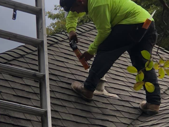 Roofer applying sealant to a vent on a shingled roof, next to a ladder.
