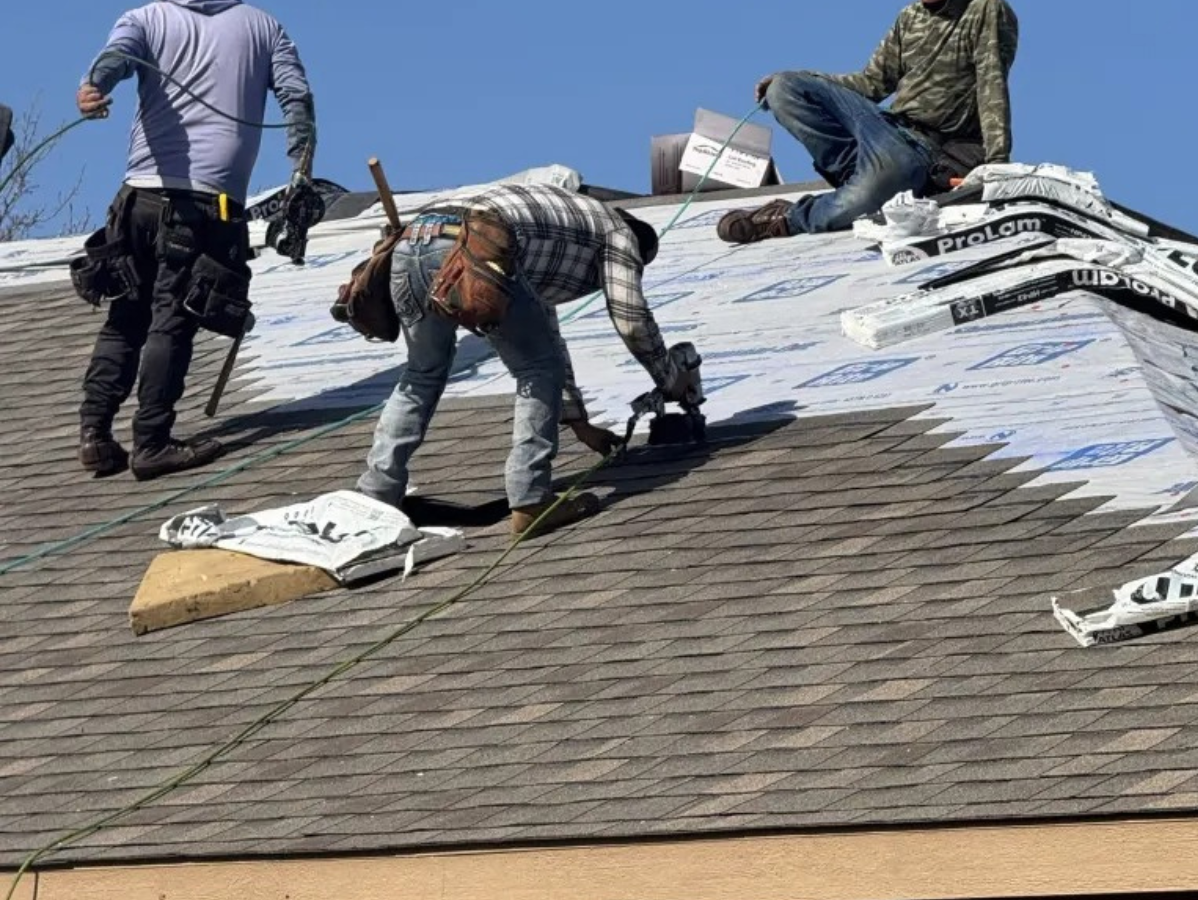 Construction workers installing shingles on a roof. Blue sky, tools, and materials are visible.