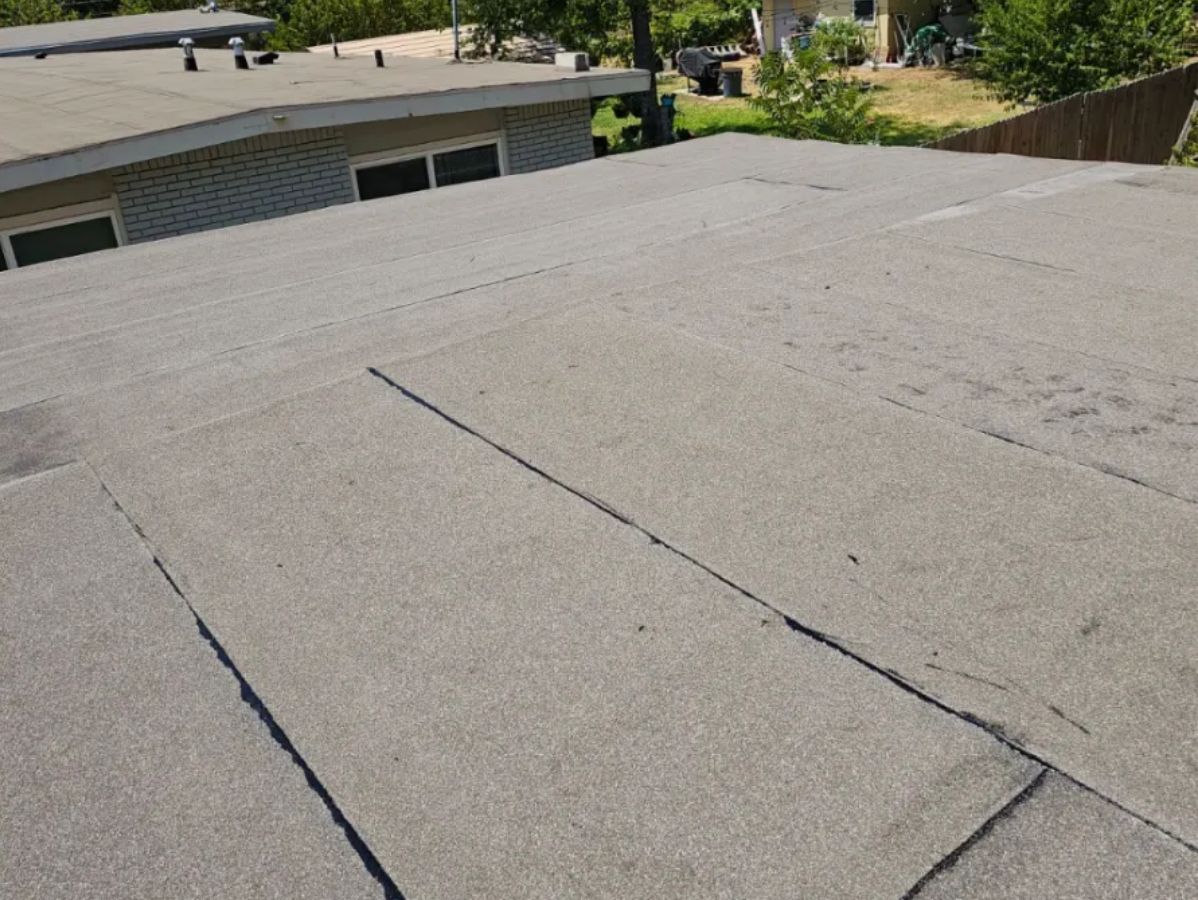 Flat, dark gray asphalt roof with visible seams. Building and trees in the background under sunlight.