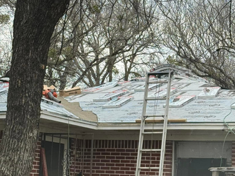 Roof repair in progress: silver ladder leans against a partially shingled roof, trees in the background.