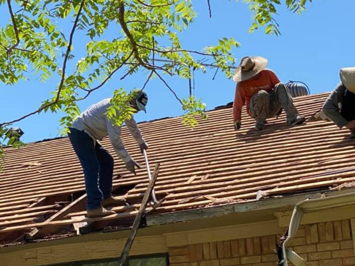 Roofers working on a house roof on a sunny day. One removes old shingles, two others crouch nearby.