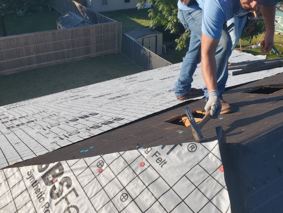 Roofer laying synthetic roofing underlayment on a dark shingled roof, outdoors.
