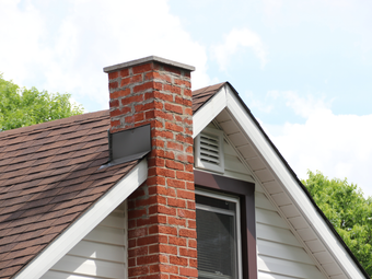Brick chimney on a brown shingled roof, next to a white house with a window and vent, against a cloudy sky.