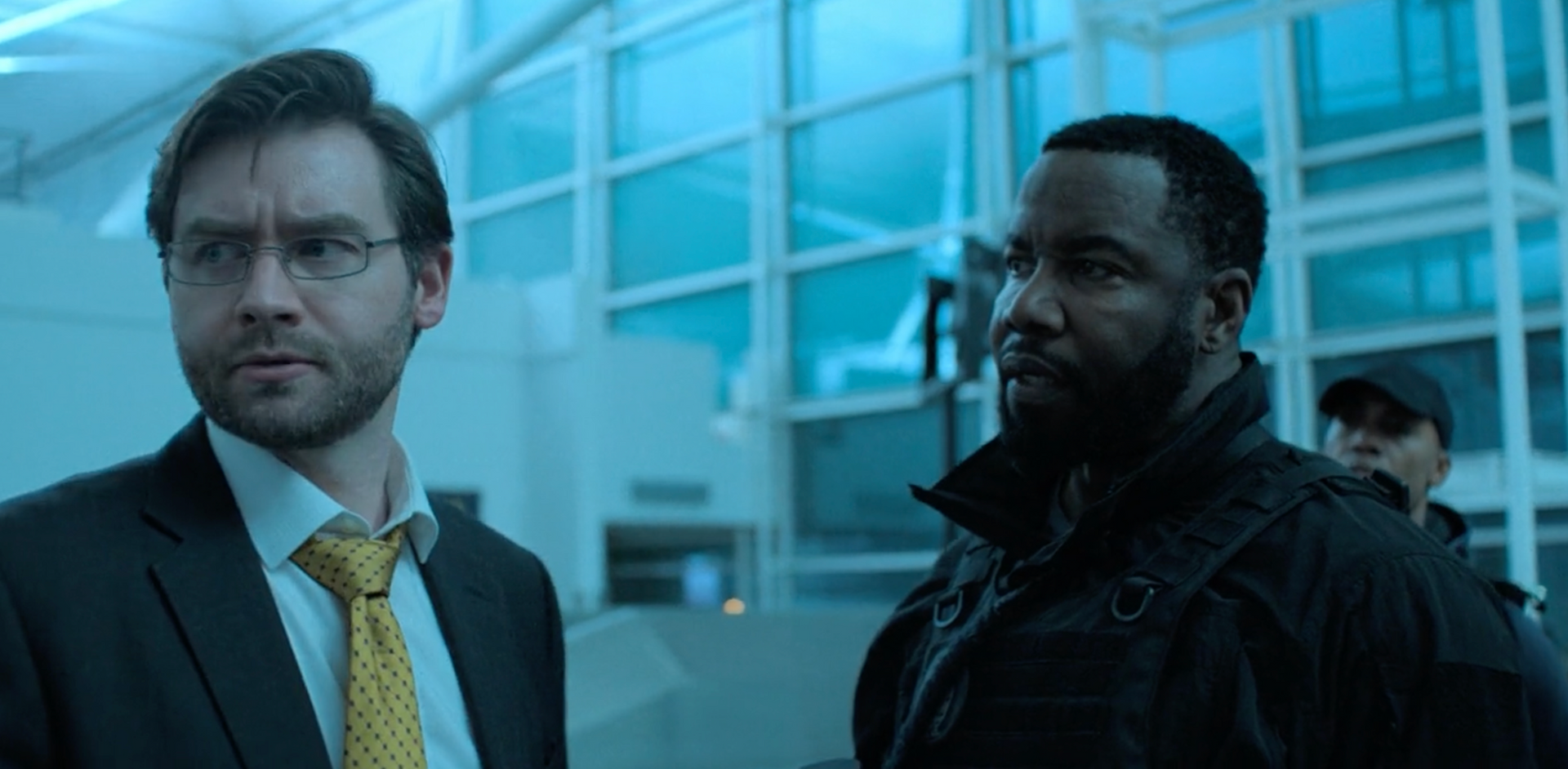 Two men in a bright, blue-lit interior. One, in a suit, looks concerned; the other, in black, looks toward the right with a neutral expression.