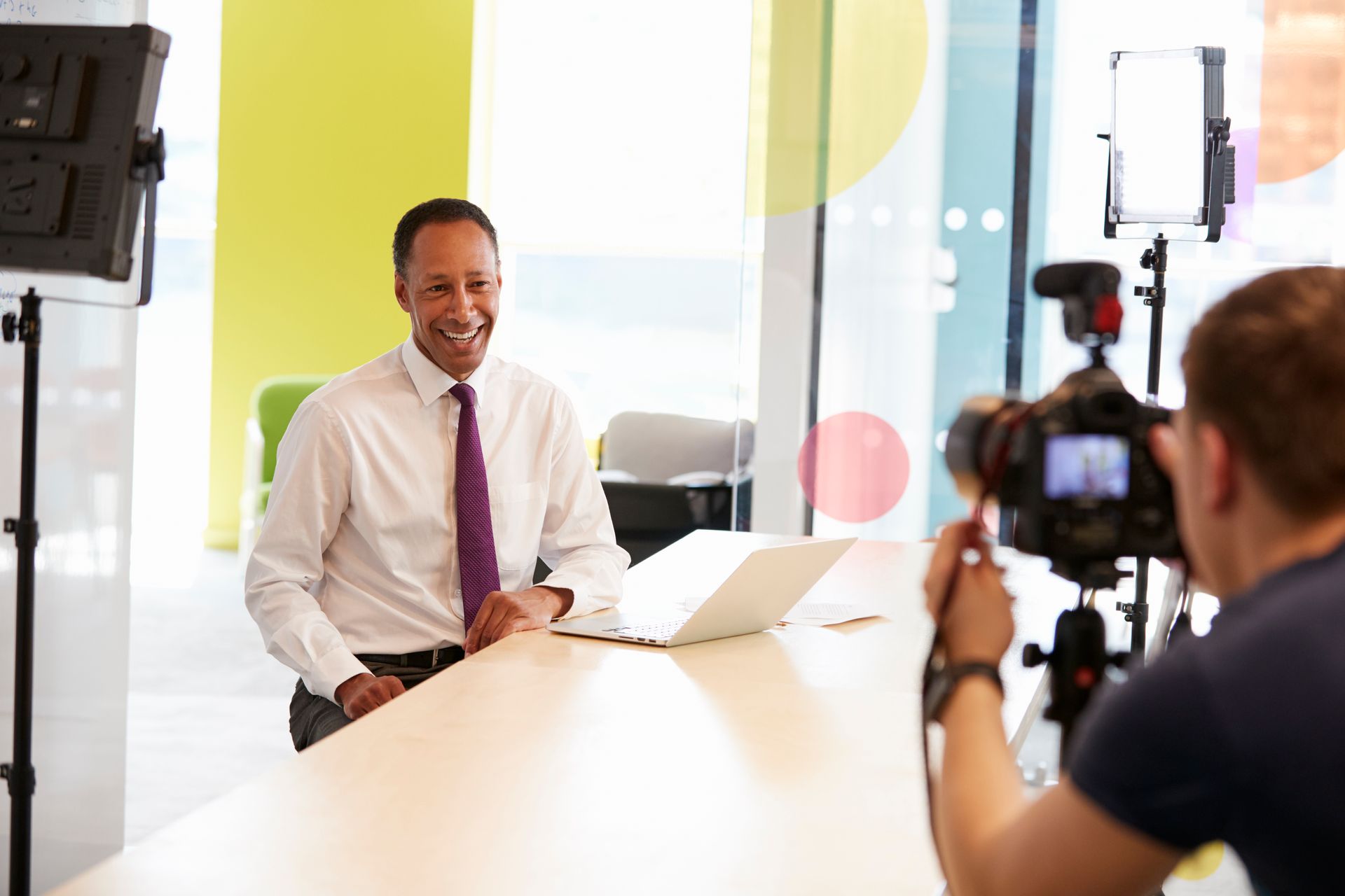 Man in a shirt and tie smiles, being filmed at a table in a modern office.