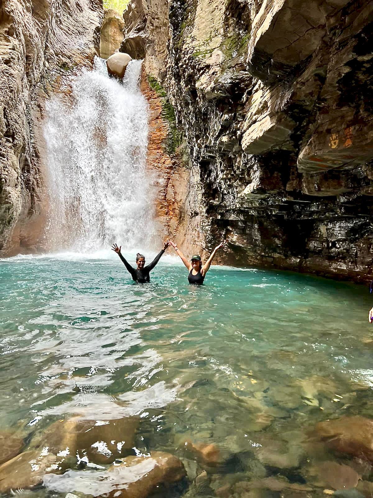 Two people swim in a turquoise pool beneath a waterfall, raising their arms. Cliffs surround the water.