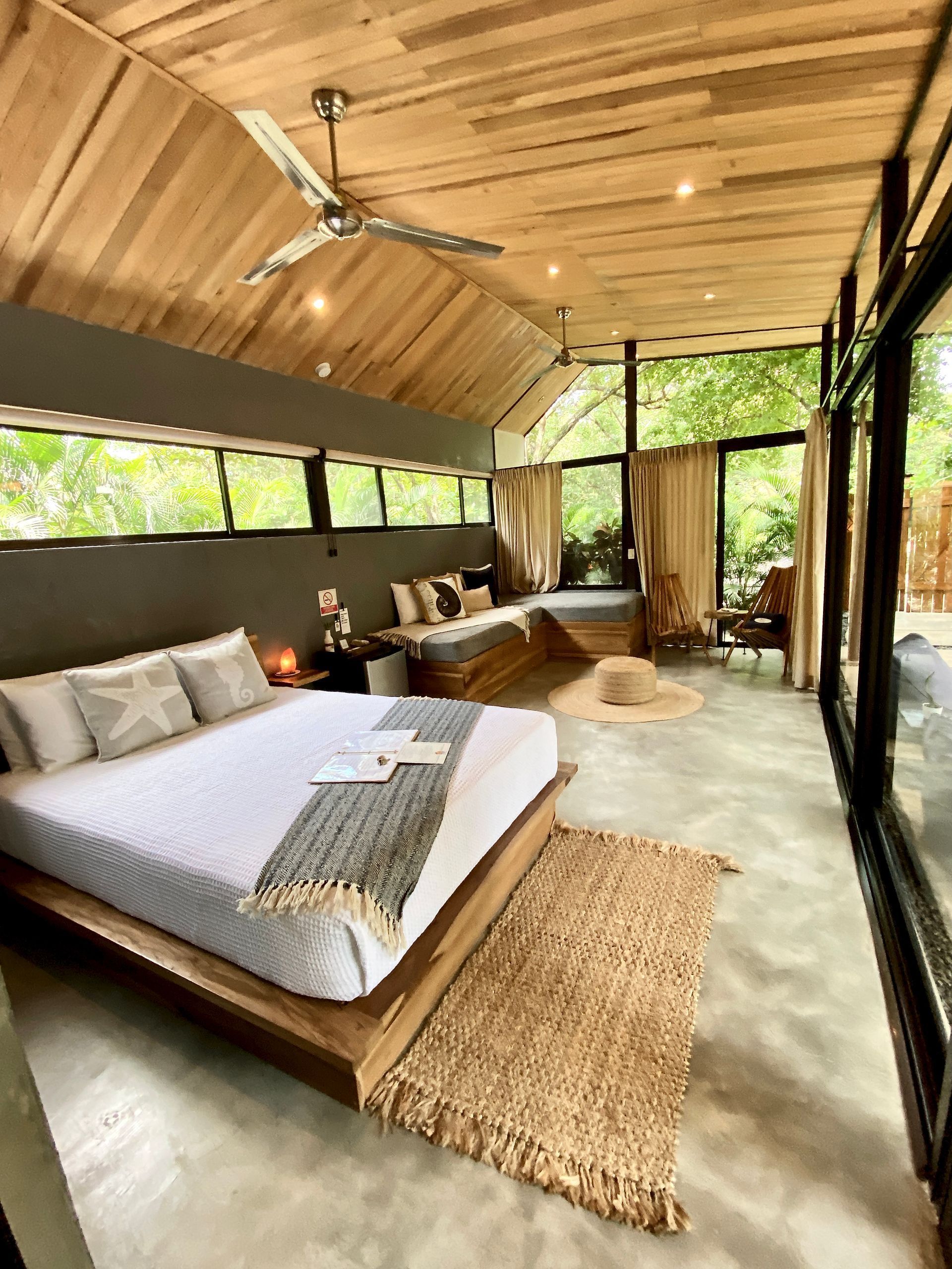 Modern bedroom with natural light, wooden ceiling and furniture, and glass walls overlooking greenery.