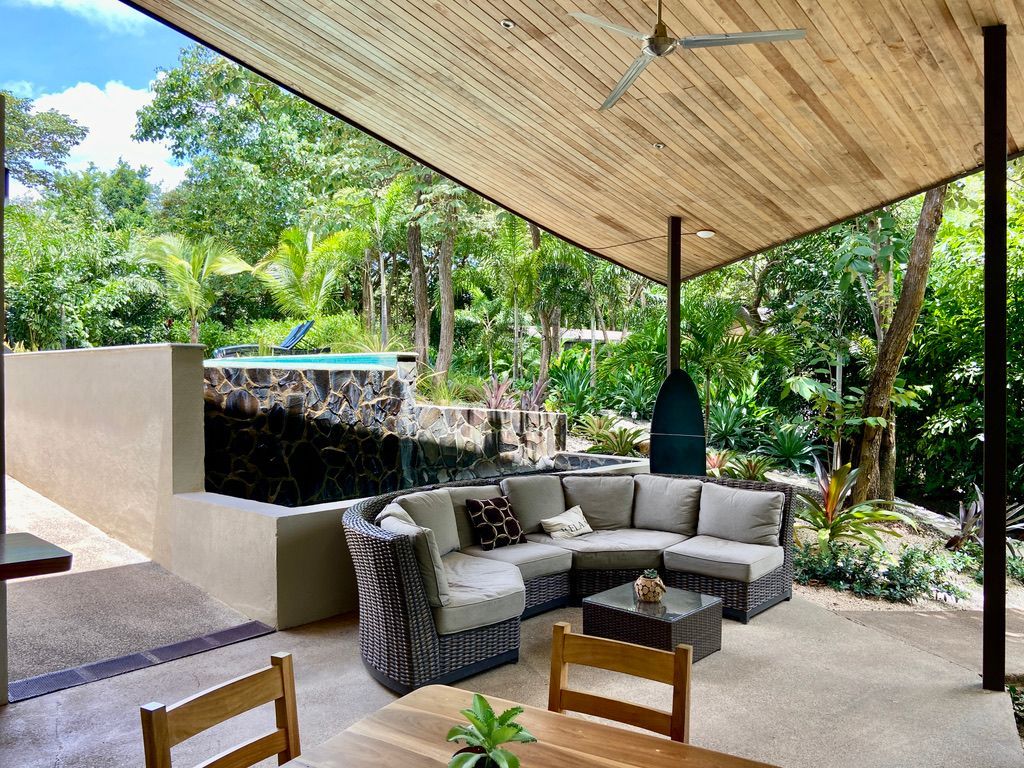 Outdoor patio with sectional sofa, table, and pool view in lush green setting.