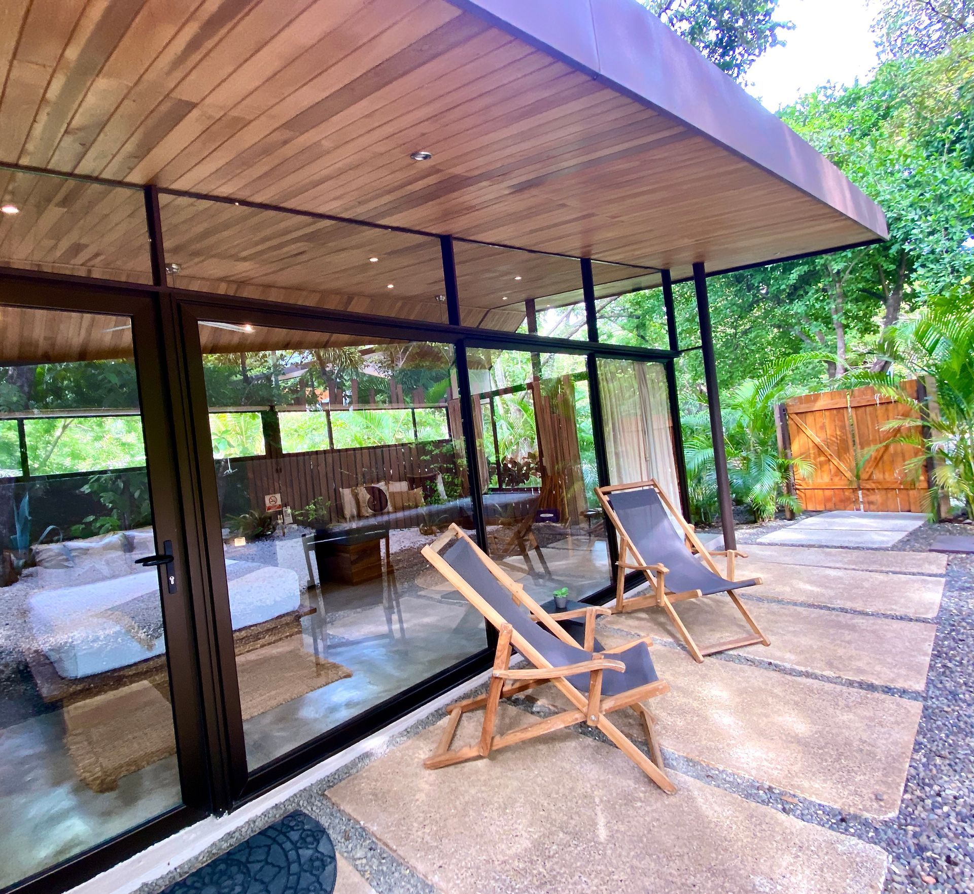 Two wooden lounge chairs sit on a patio outside a glass-walled building with wooden ceiling; wooden gate in background.