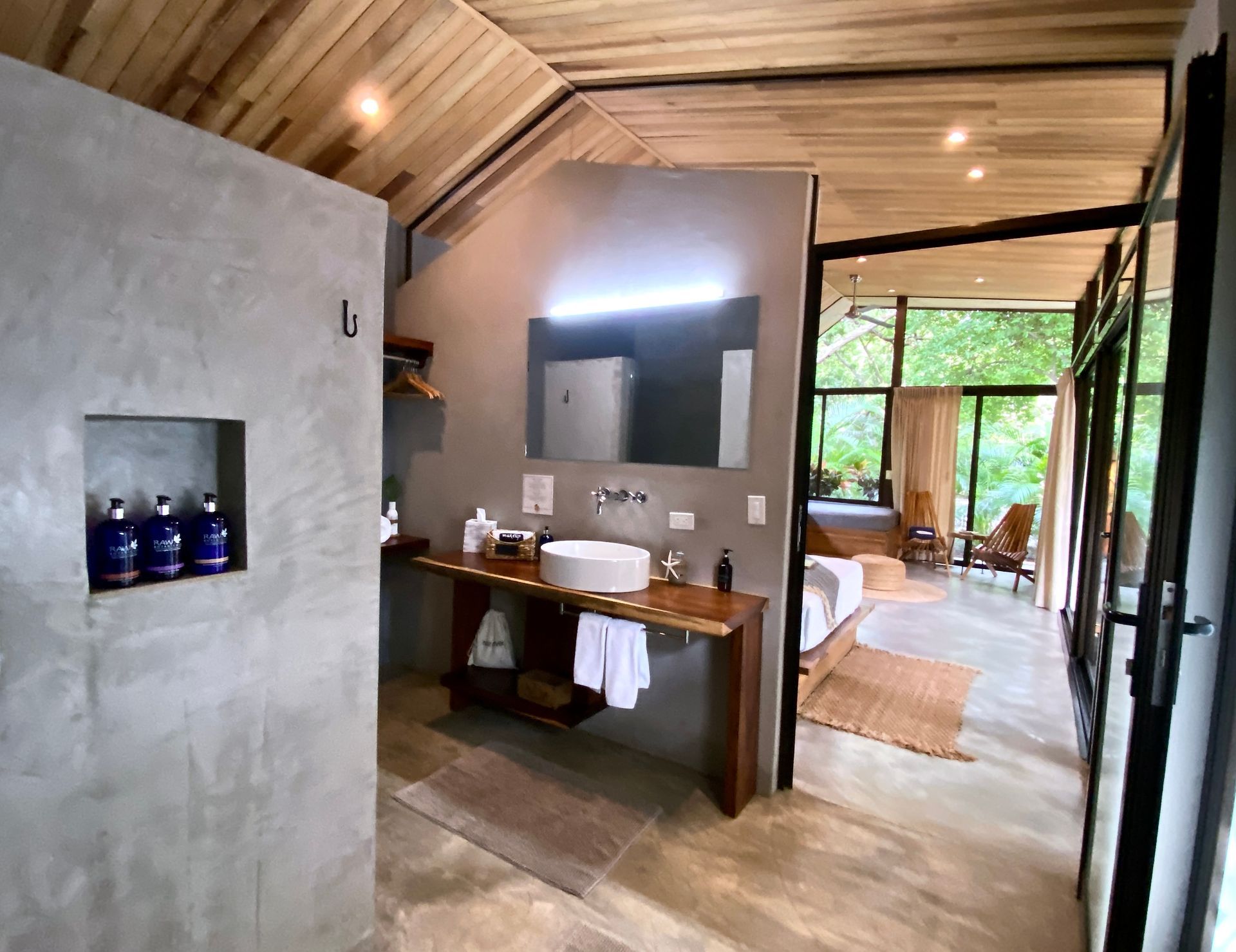 Bathroom with concrete walls, wood accents, and a view into a bedroom with large windows.