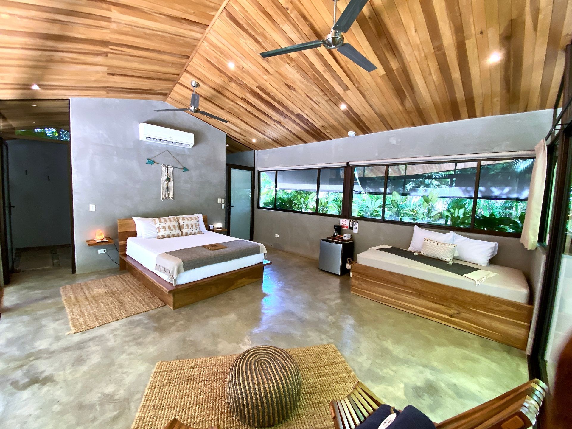 A bright modern bedroom with a wooden ceiling, bed, daybed, and large windows overlooking greenery.