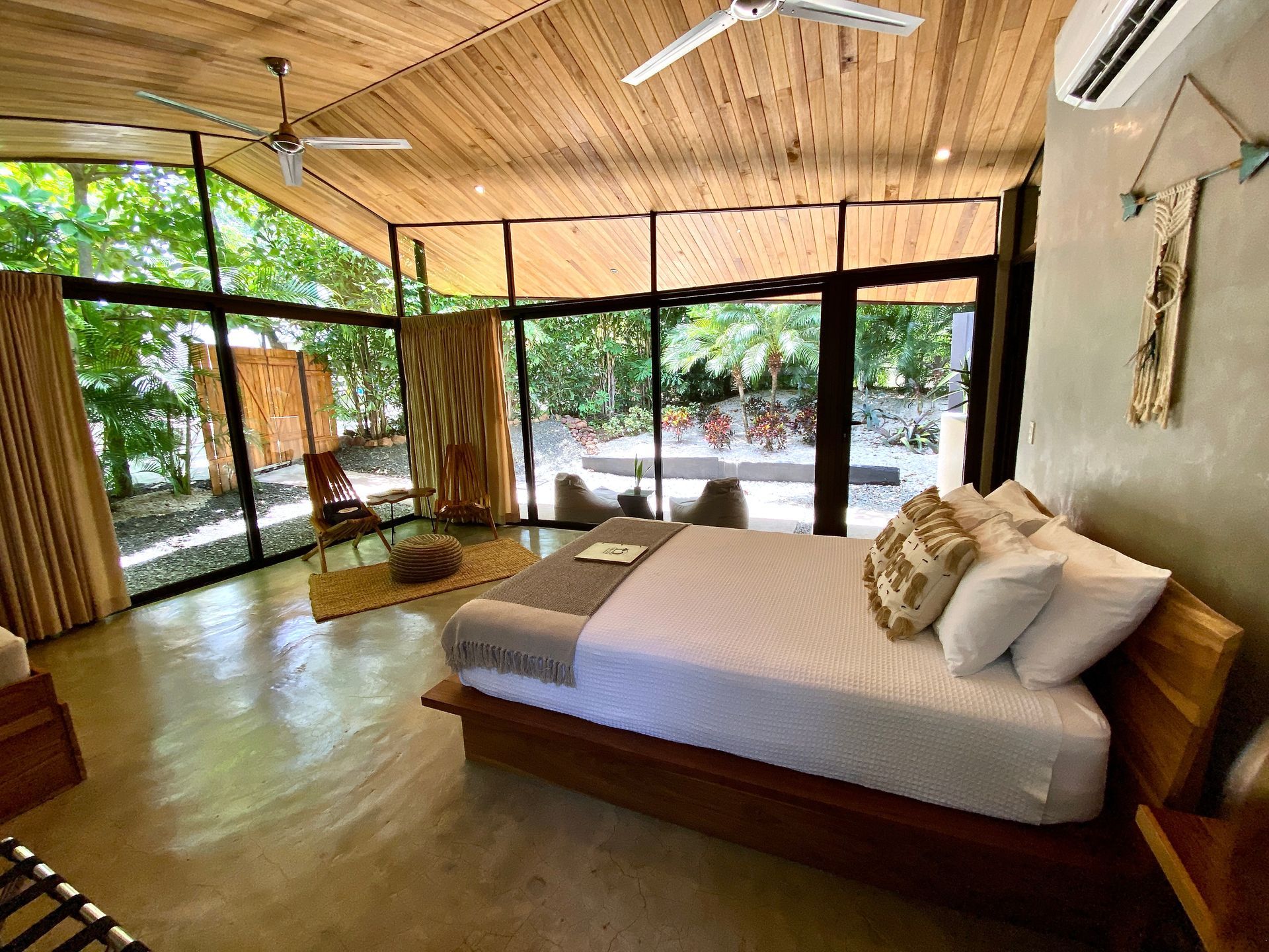 Bedroom with large windows overlooking lush greenery. Wooden bed, ceiling fans, and bamboo accents.
