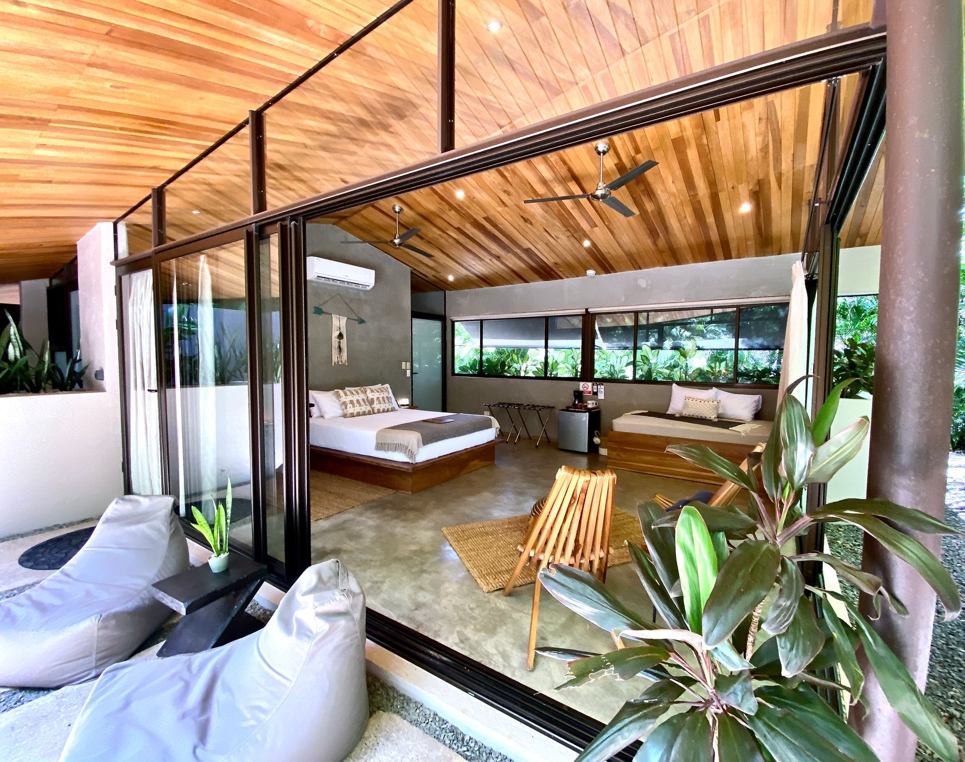 Modern open-plan room with sliding glass doors, seating areas, a bed, and a view of lush greenery.