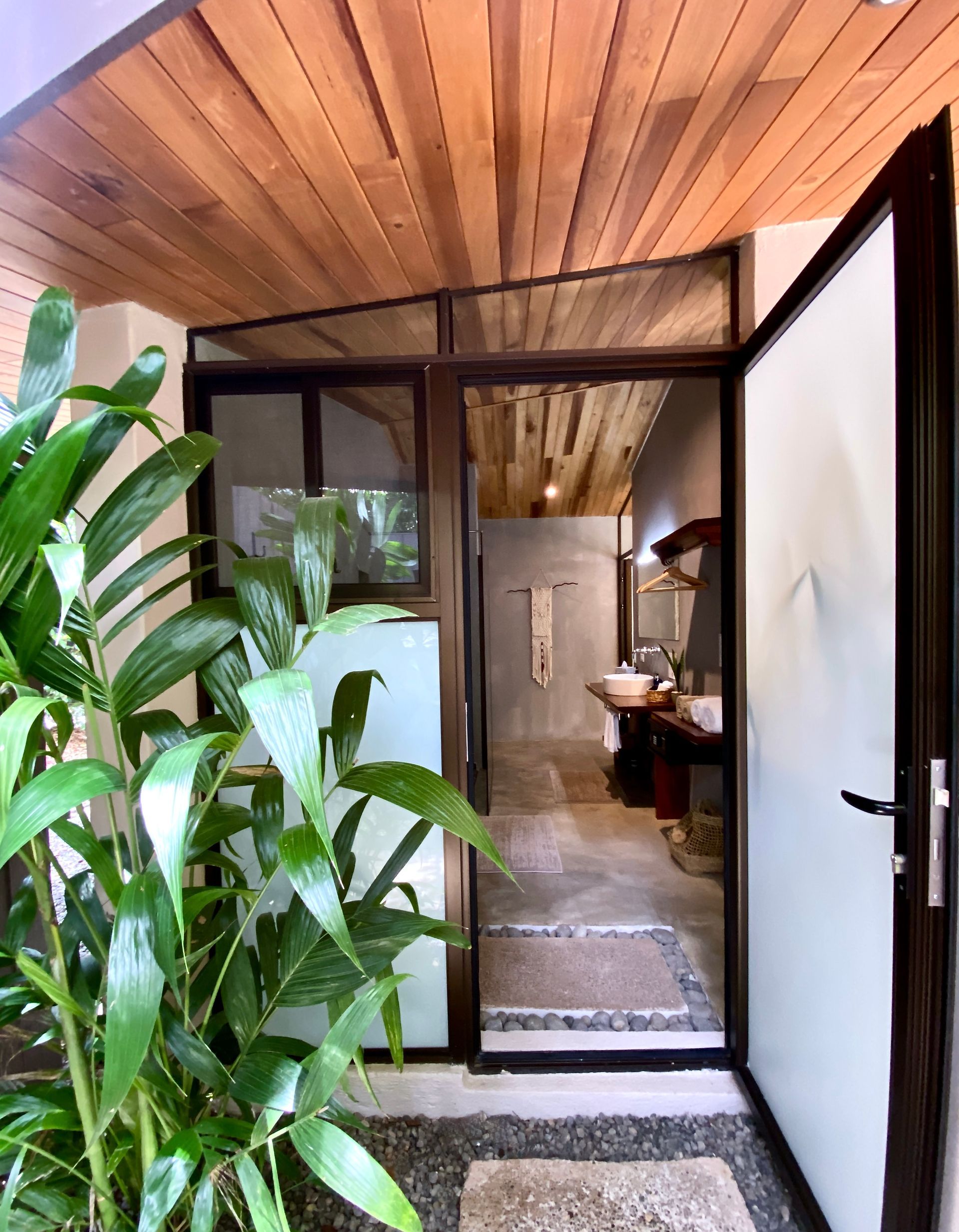 Entryway to a bathroom, lush green plants frame open door. Light wood ceiling, grey stone floor, vanity visible.