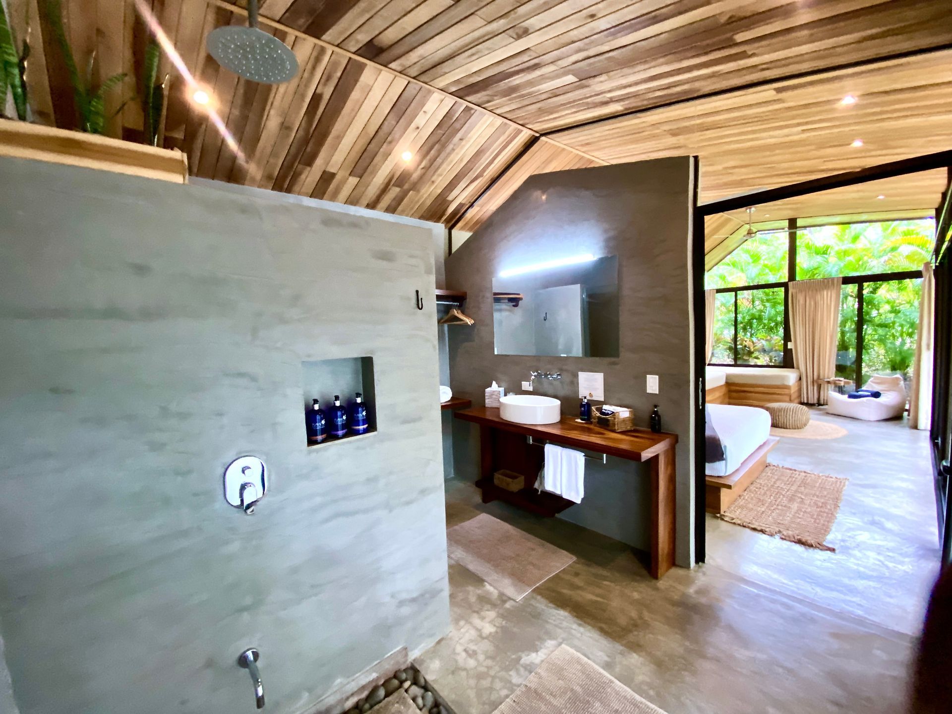 Bathroom with open shower and sink, connected to bedroom with natural light and wooden accents.