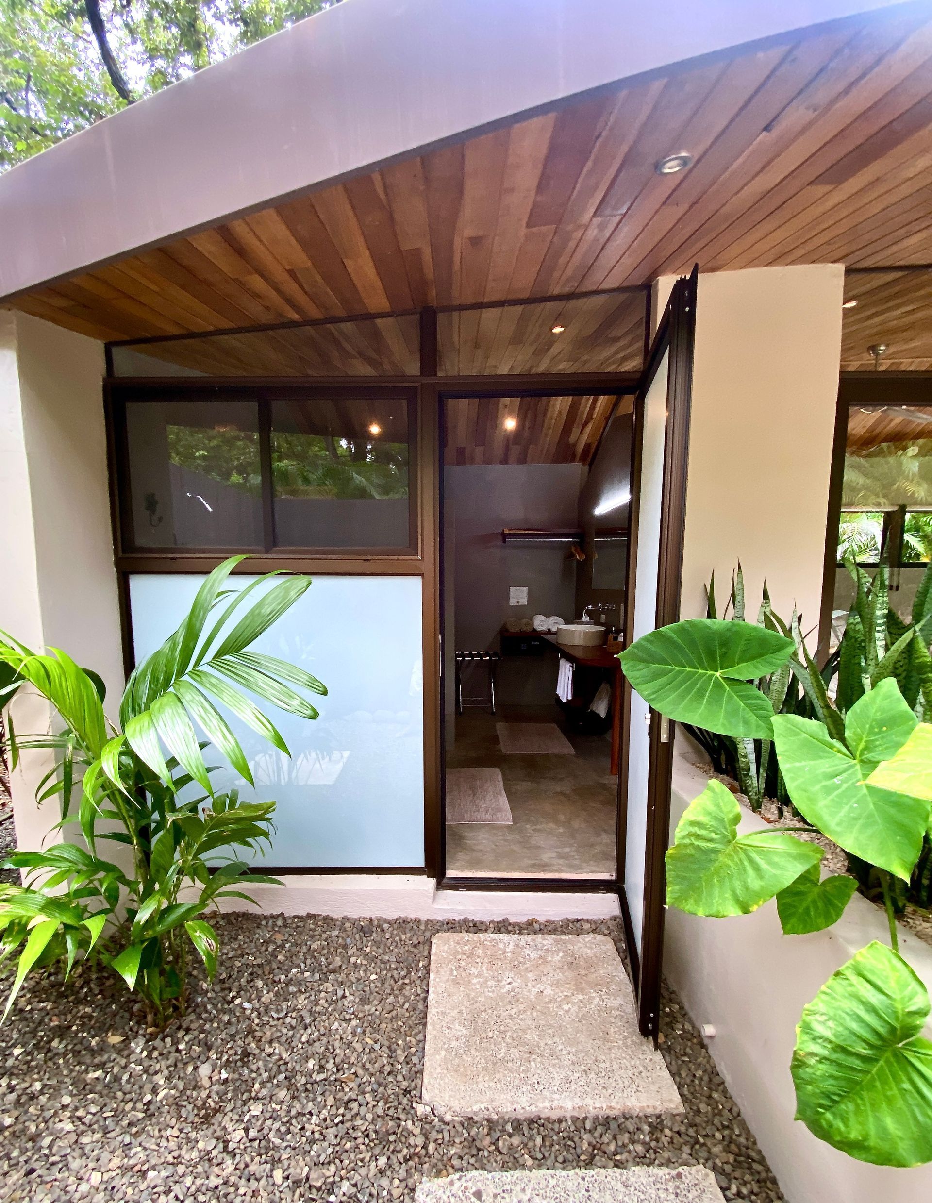 Entryway with open door to a bathroom, framed by wood and plants. Gravel path leads to the entrance.