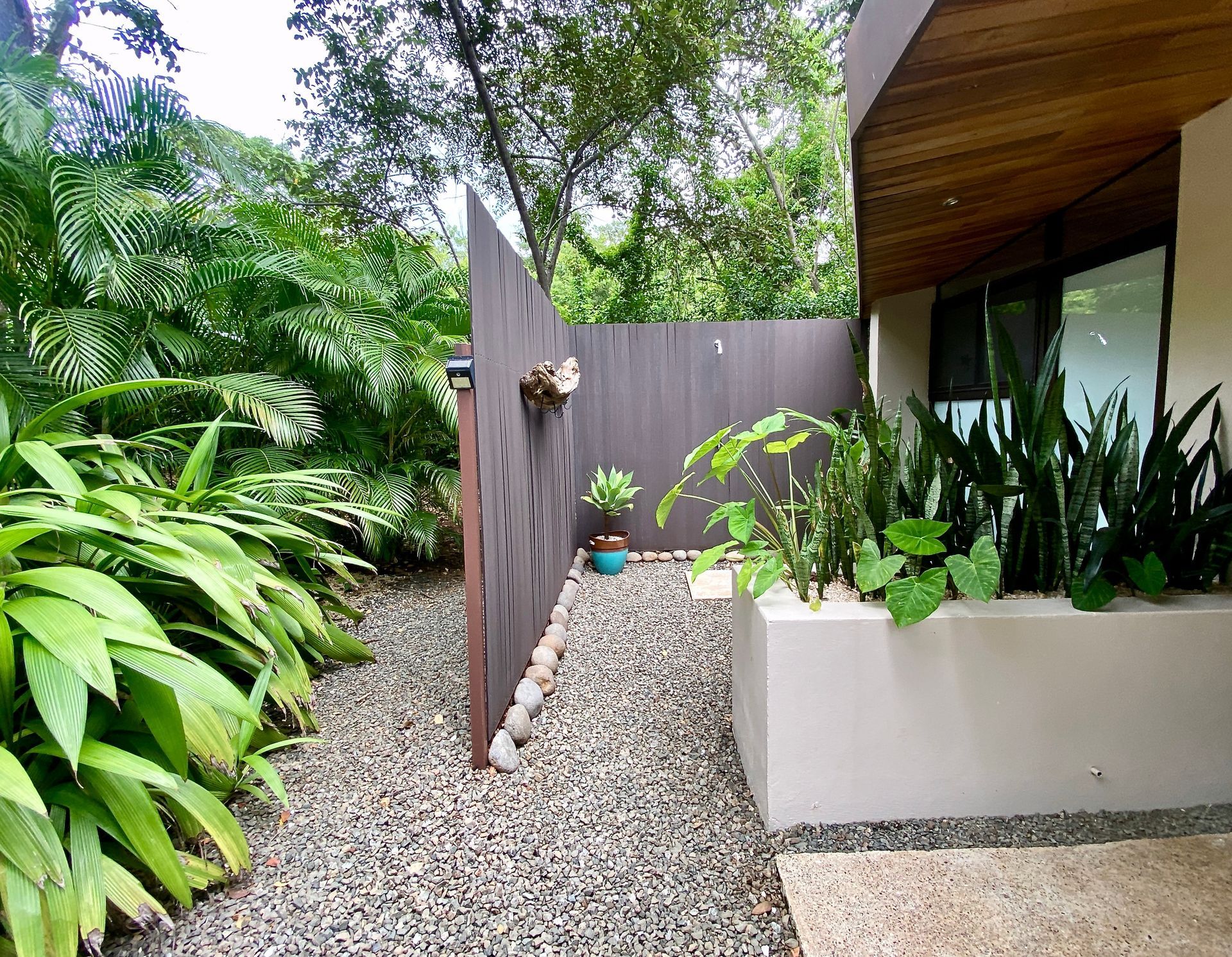 Outdoor shower area with a gravel path, surrounded by lush greenery and wood paneling.