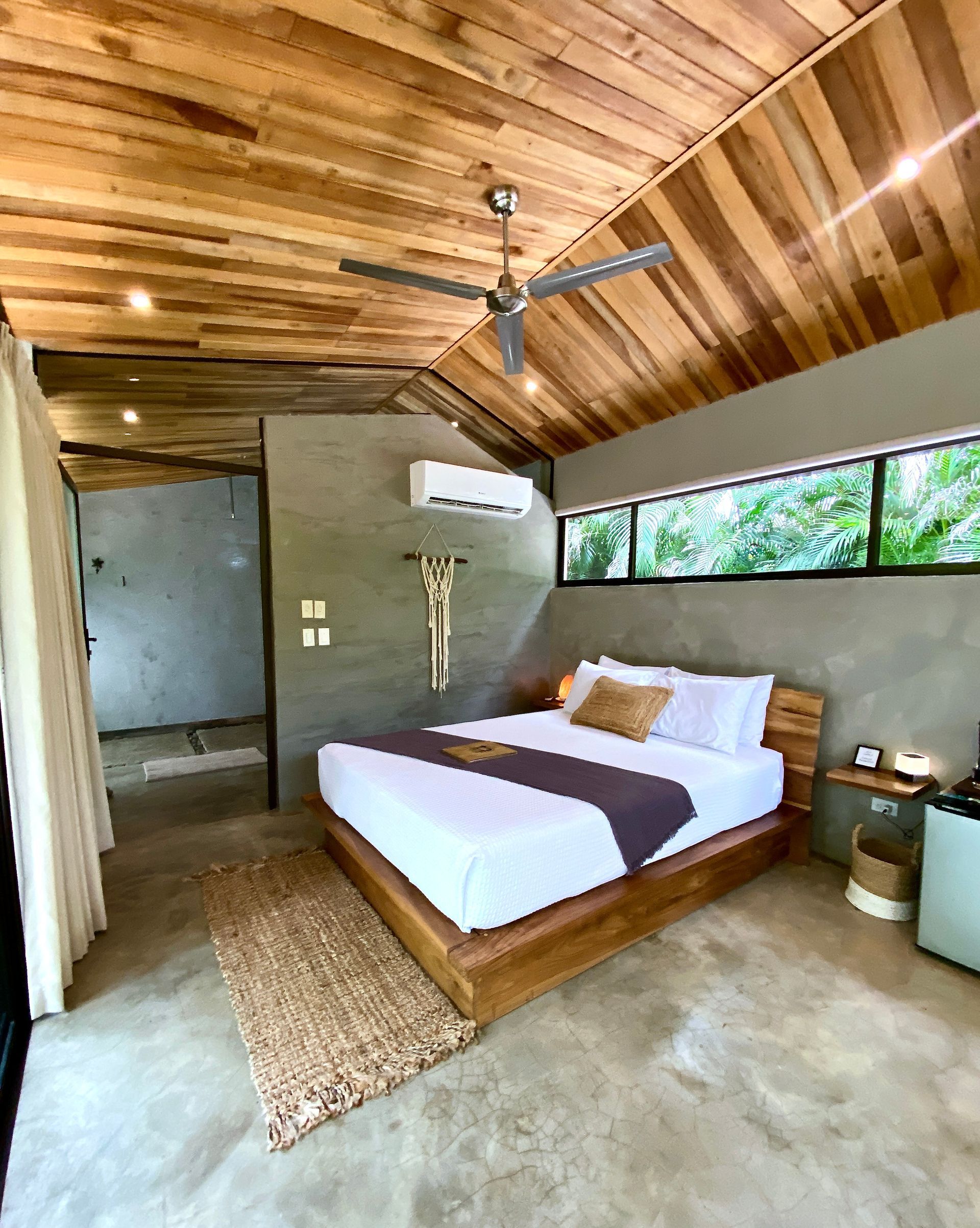 Bedroom with wood ceiling, bed, and window overlooking green foliage.