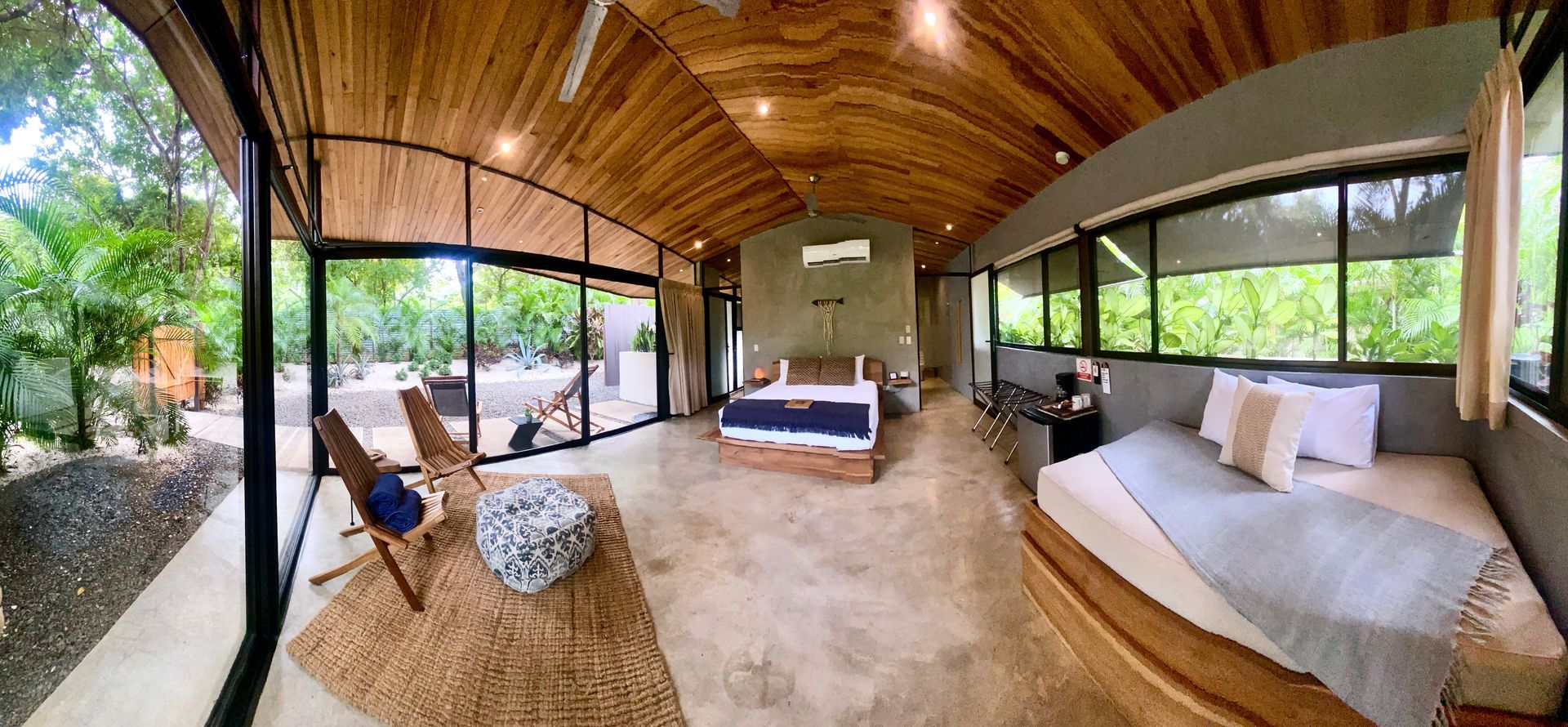 A modern bedroom with floor-to-ceiling windows and wooden ceiling; a bed with white and blue linens and a sofa.
