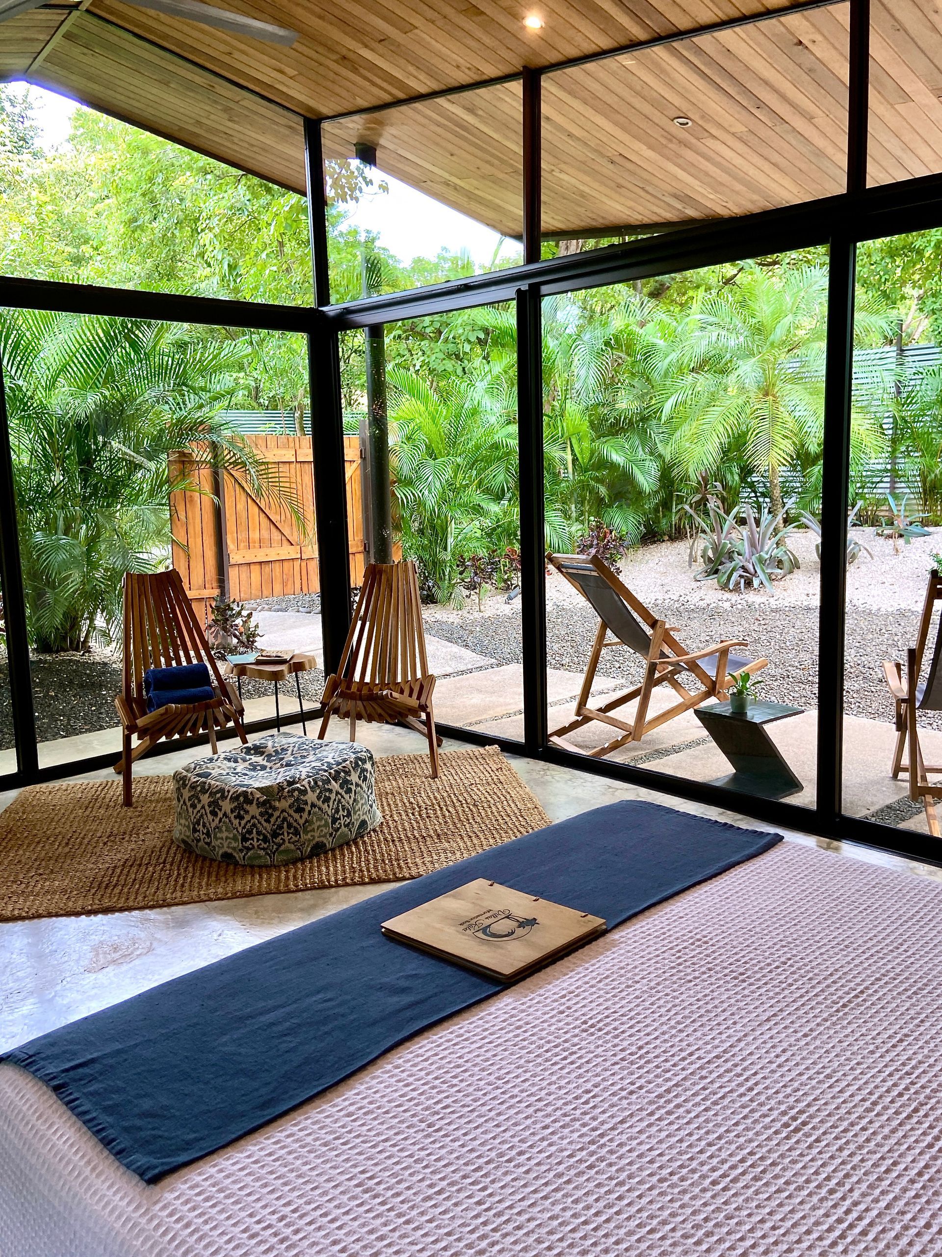 Glass-walled room with yoga mat, chairs, and outdoor view; wooden ceiling.