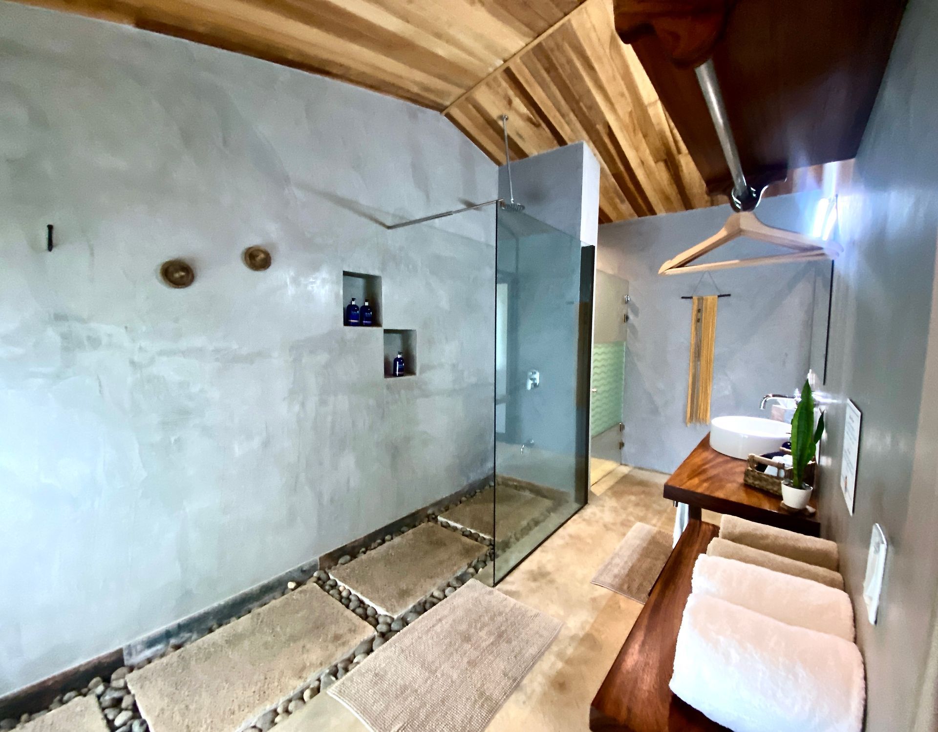 Bathroom with gray concrete walls, wooden ceiling, glass shower, and stone floor mats.