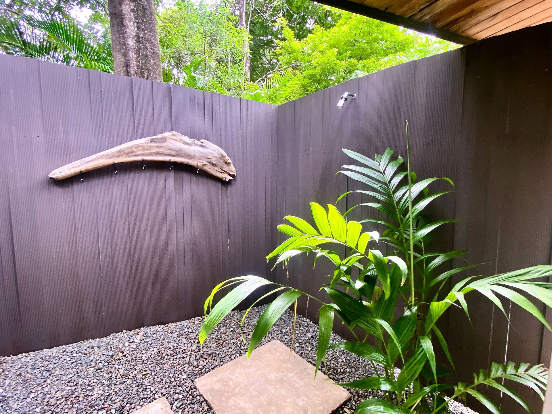 Outdoor shower with brown walls, driftwood, green plants, and stone floor.
