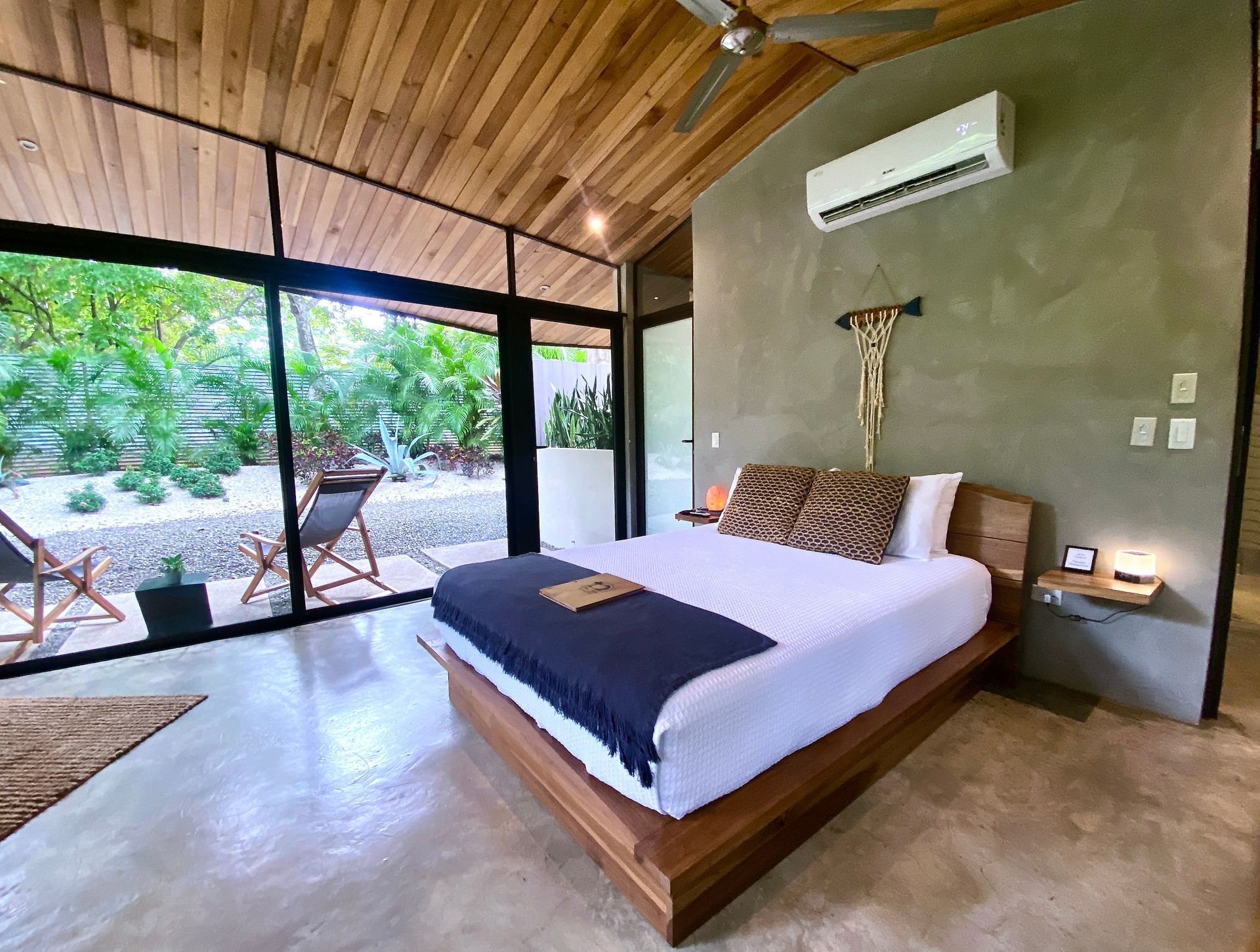 Bedroom with wooden bed, large windows, garden view, concrete floor, and air conditioner.