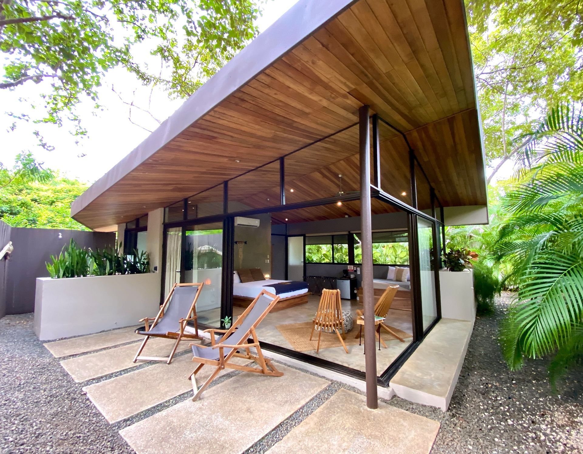 Modern cabin with glass walls, wooden ceiling, patio with chairs. Lush greenery surrounds.