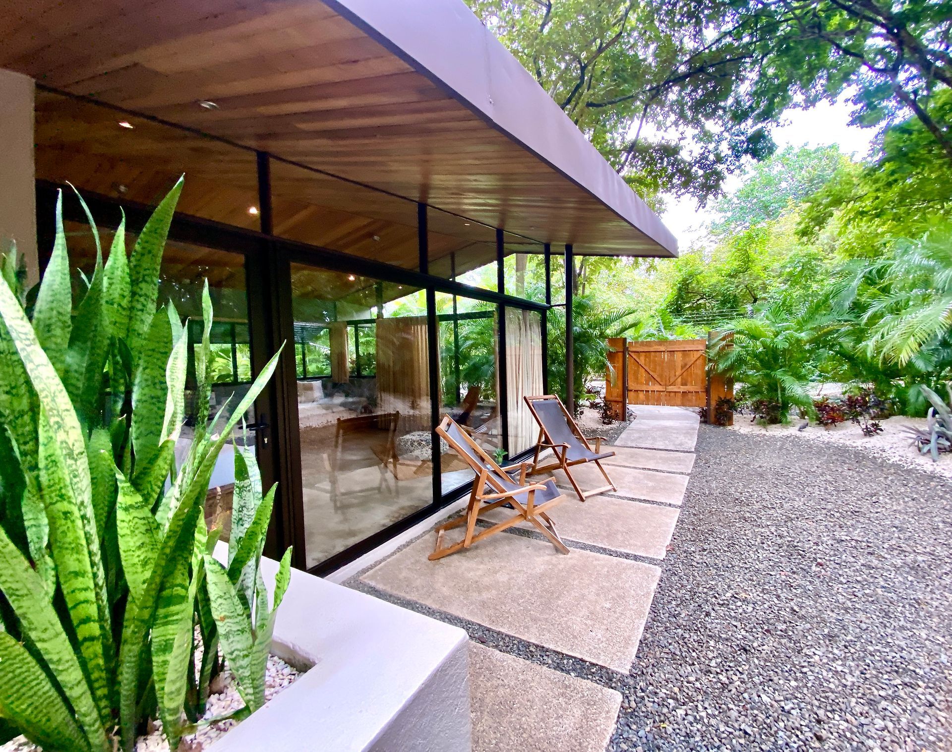 Modern home exterior with floor-to-ceiling windows, wooden ceiling, lounge chairs, and lush greenery.