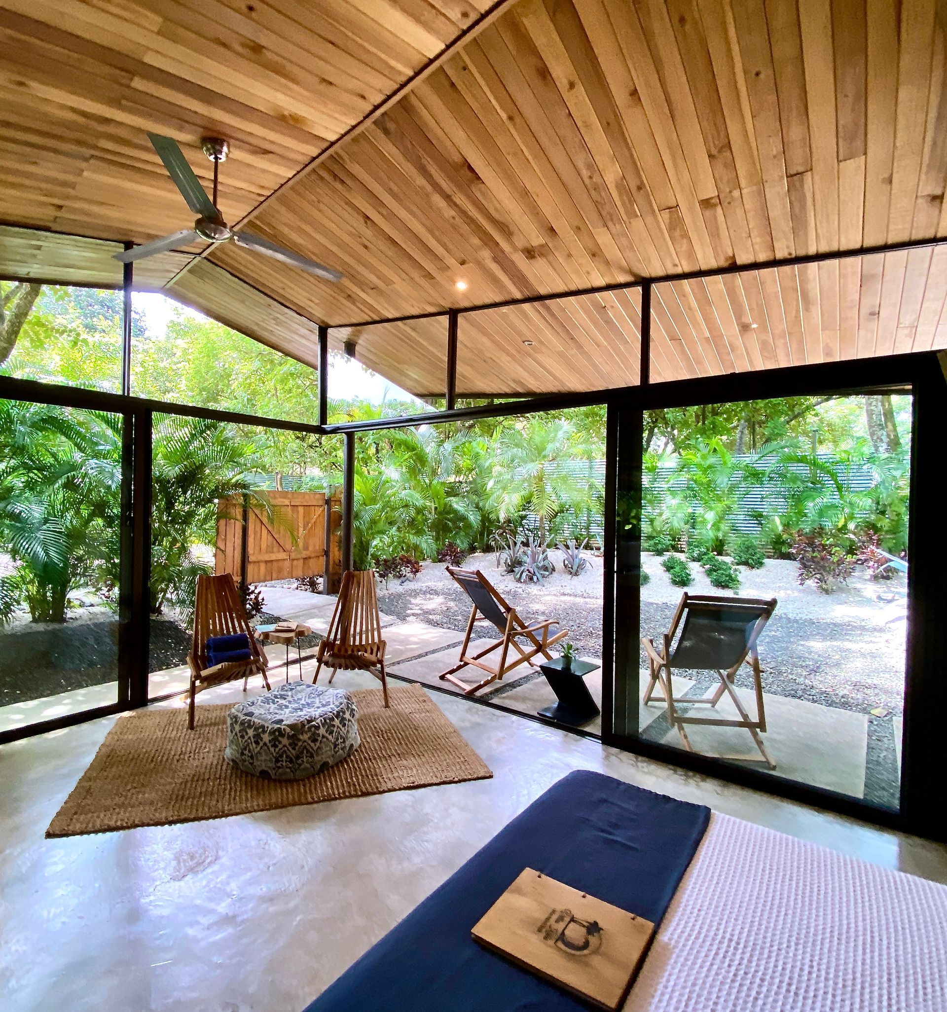A light-filled room with wooden ceiling and glass walls opening to a lush garden, featuring seating and a bed.