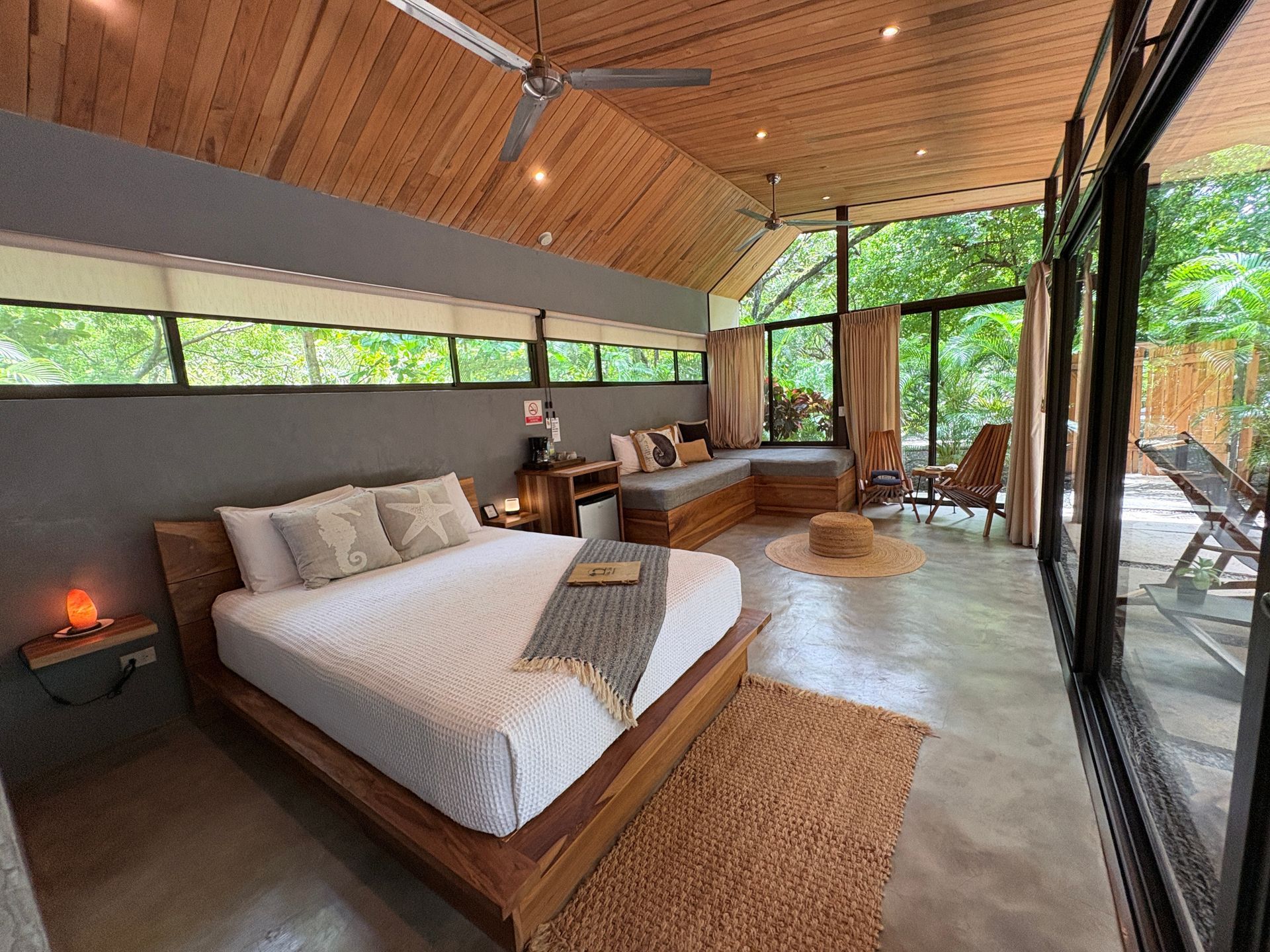 Bedroom with a wooden bed, large windows, and a seating area, surrounded by trees.